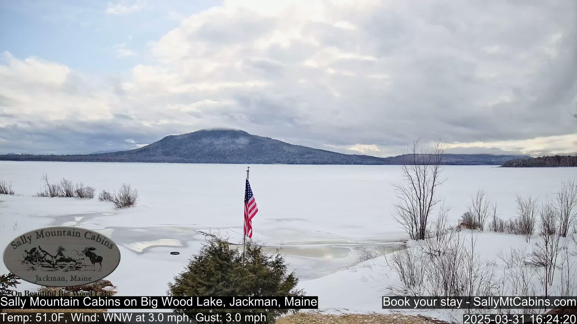 A snow-covered frozen lake stretches out before a mountain range under a cloudy sky, with an American flag planted in the foreground.