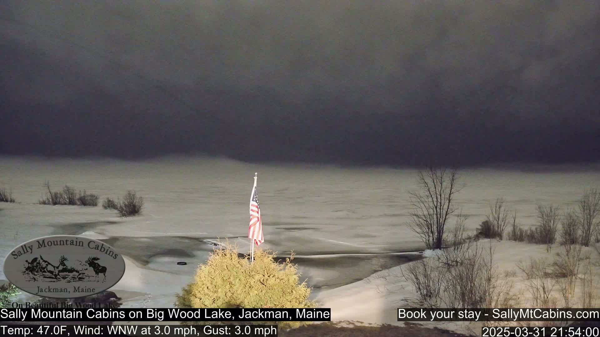 A snow-covered landscape with a frozen lake under a dark, overcast sky, and an American flag on a pole in the foreground.