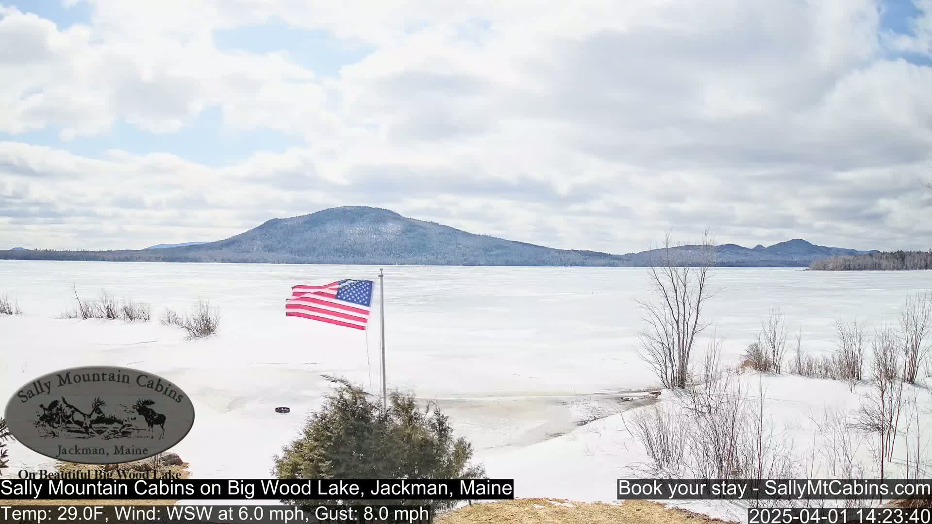 A partially frozen lake stretches to snow-covered mountains under a partly cloudy sky, with an American flag waving in the foreground.
