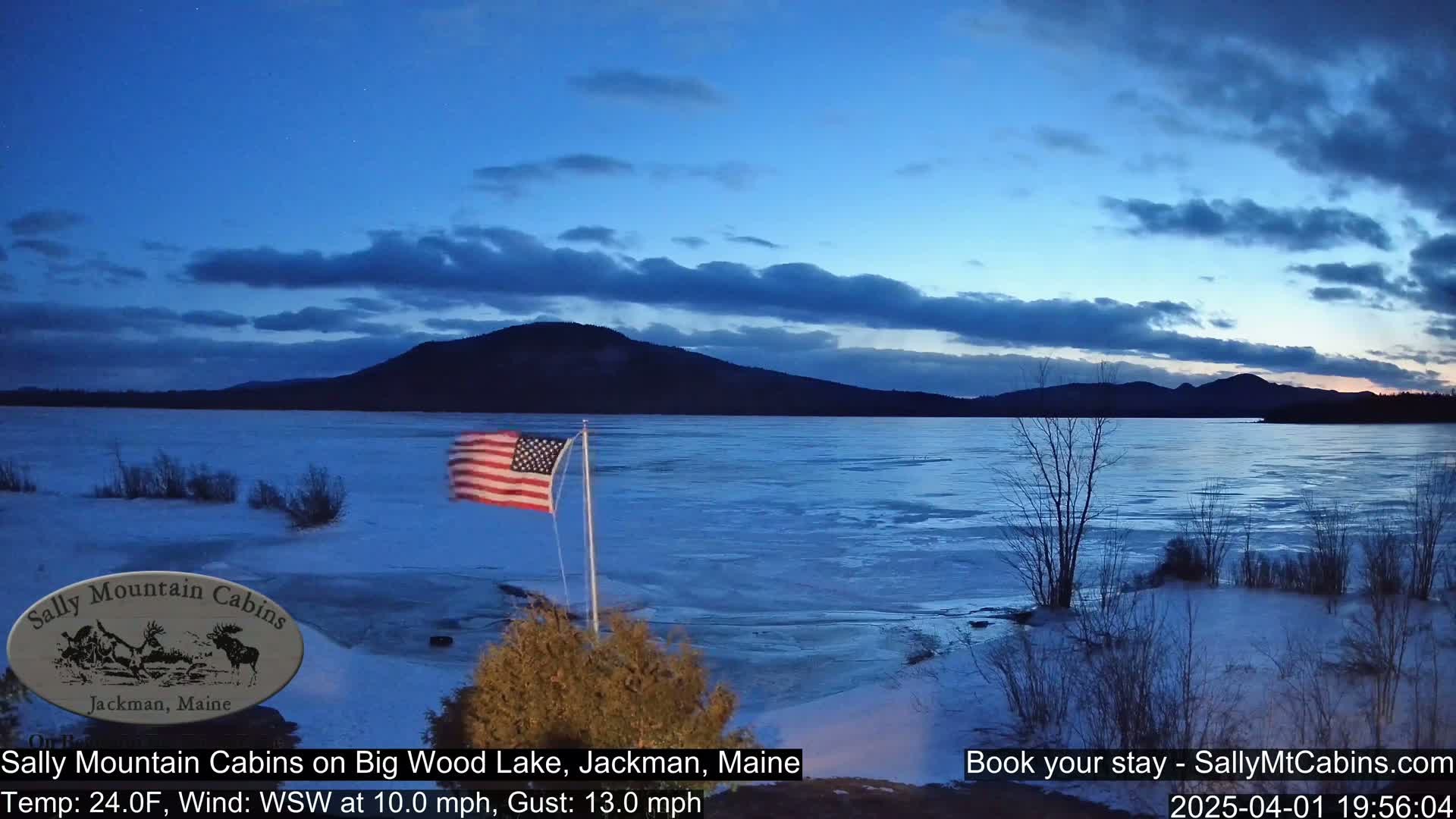 An American flag flies near a partially frozen lake at dusk, with a mountain range visible in the background under a partly cloudy sky.