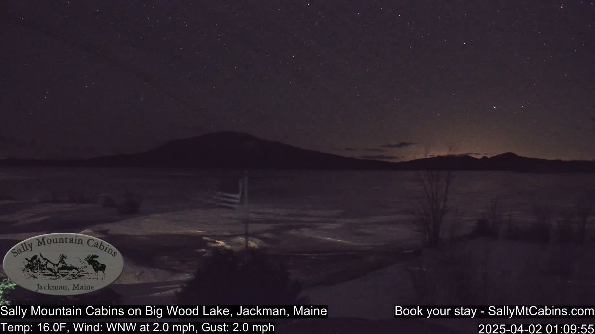 A nighttime view of a dark, partially frozen lake under a starry sky, with a distant mountain range silhouetted against the horizon.