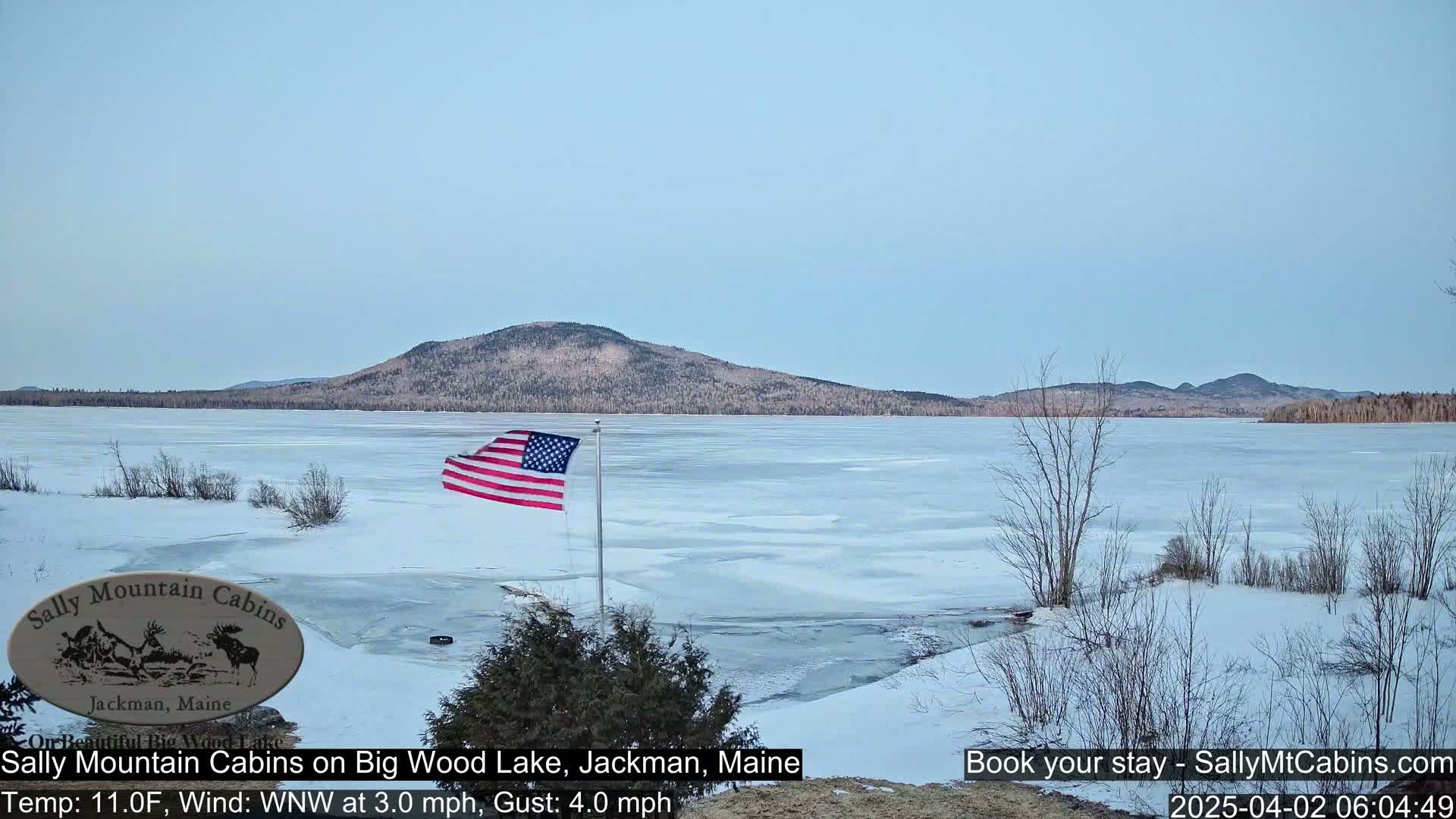 A partially frozen lake stretches to a snow-covered hill under a pale blue sky, with an American flag waving in the cold wind.