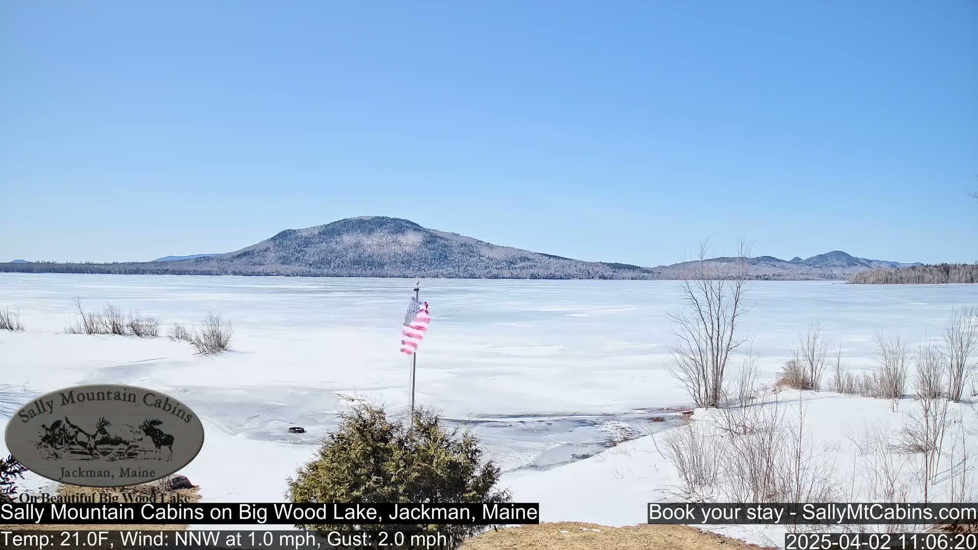 A mostly frozen lake stretches towards a snow-dusted mountain range under a clear blue sky.