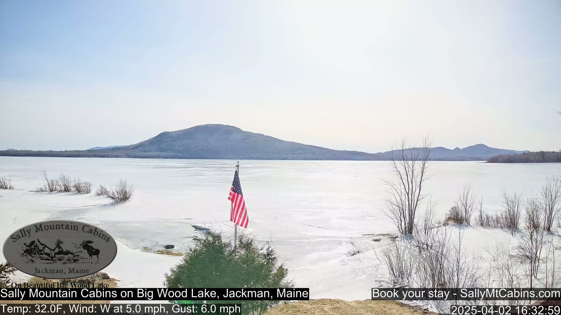 A partially frozen lake with a snow-covered shore and a distant mountain range under a bright, sunny sky, an American flag planted near the shoreline.