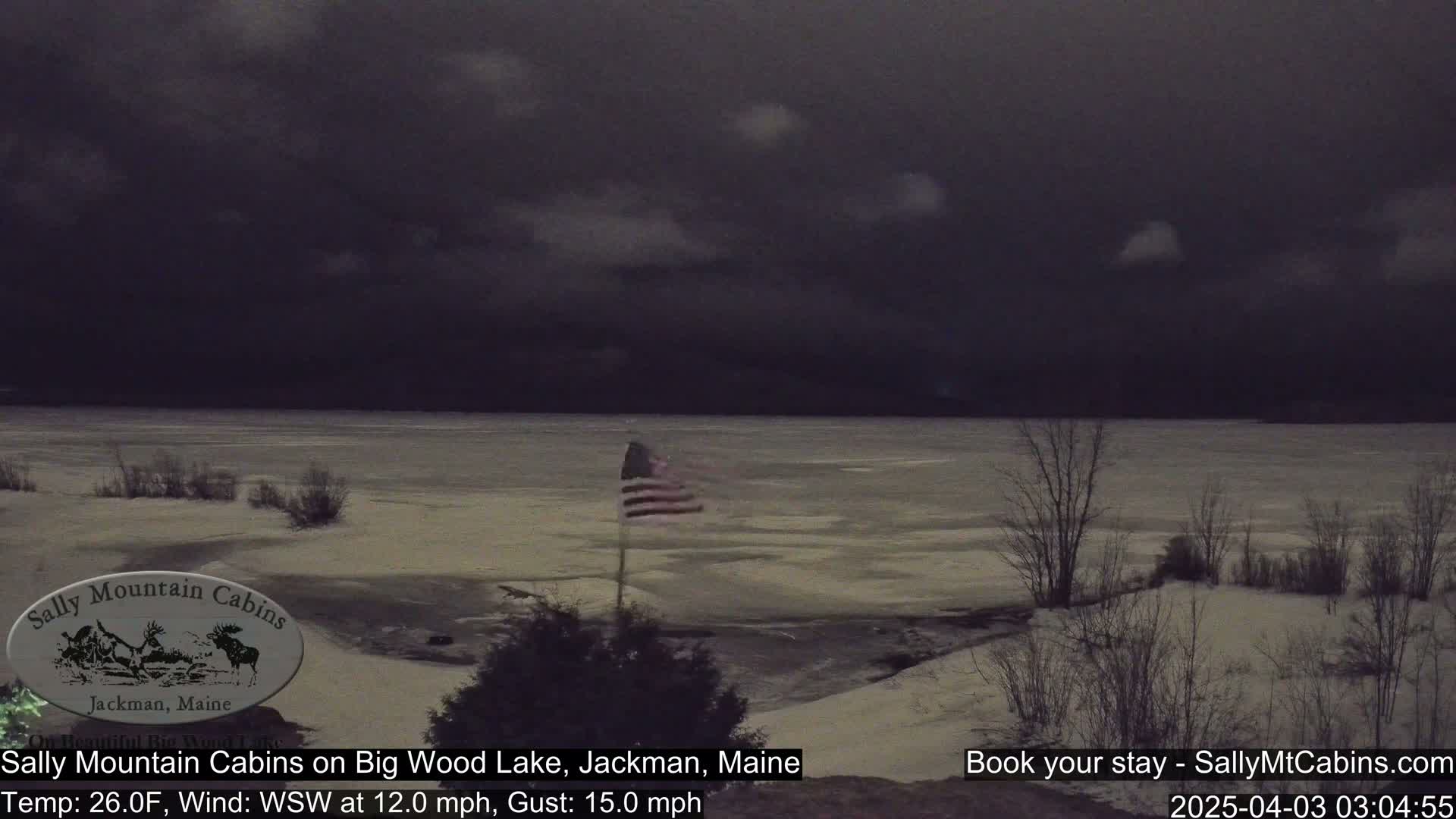 A partially frozen lake is visible under a dark, cloudy sky, with a small American flag waving near the shoreline and snow-covered land.