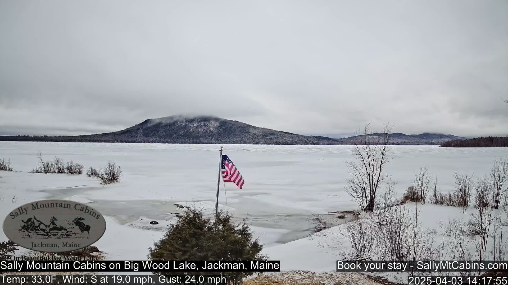 A partly cloudy, cold day shows a snow-covered frozen lake with a distant mountain range under a gray sky, and an American flag is waving near the shore.