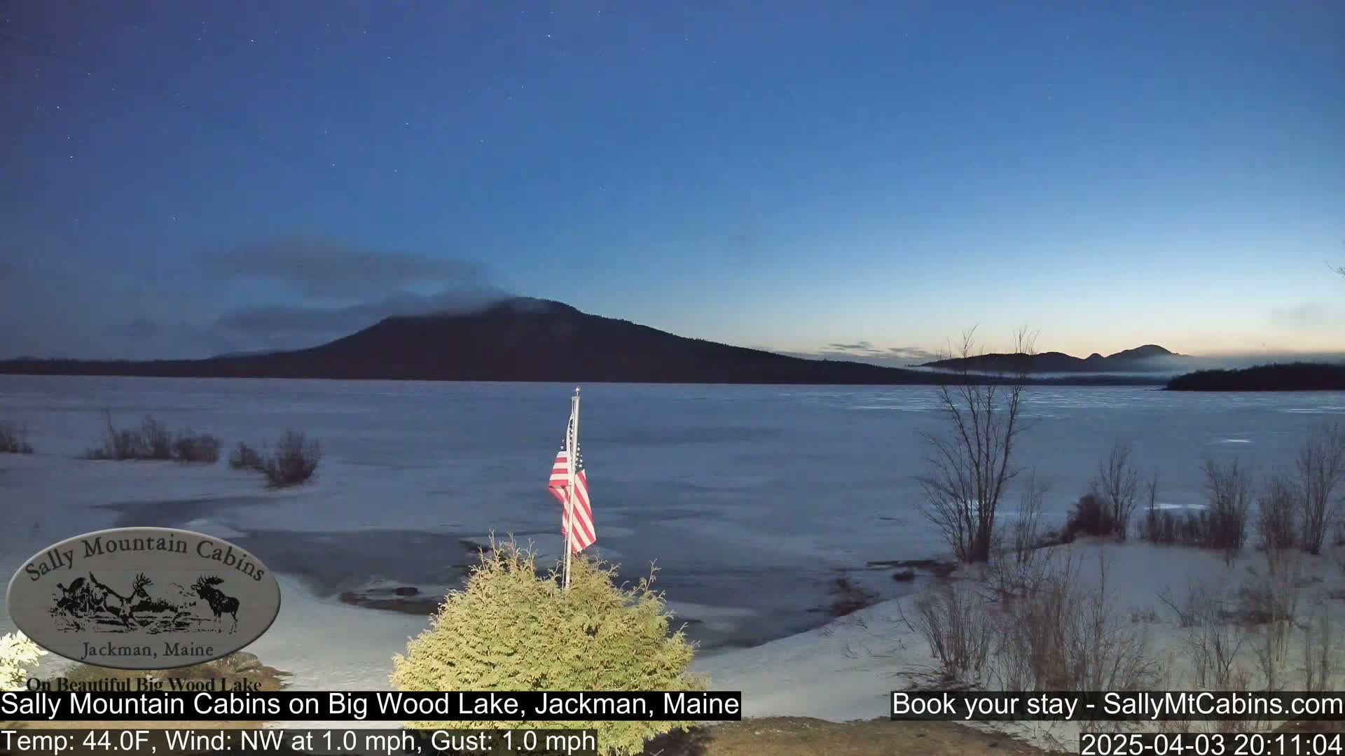 A partially frozen lake under a twilight sky, with a mountain range in the background and an American flag in the foreground.