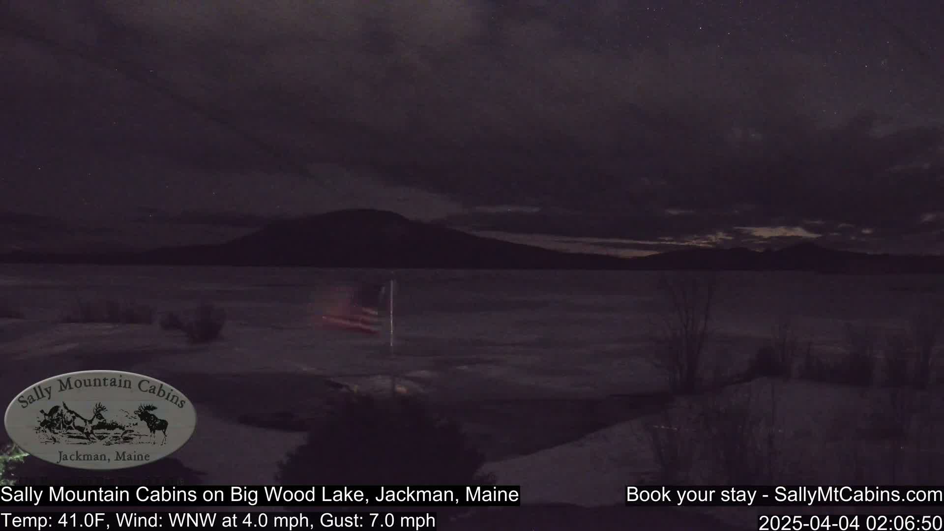 A nighttime view of a mostly calm lake under a dark, cloudy sky with a faintly visible flagpole and distant mountains.