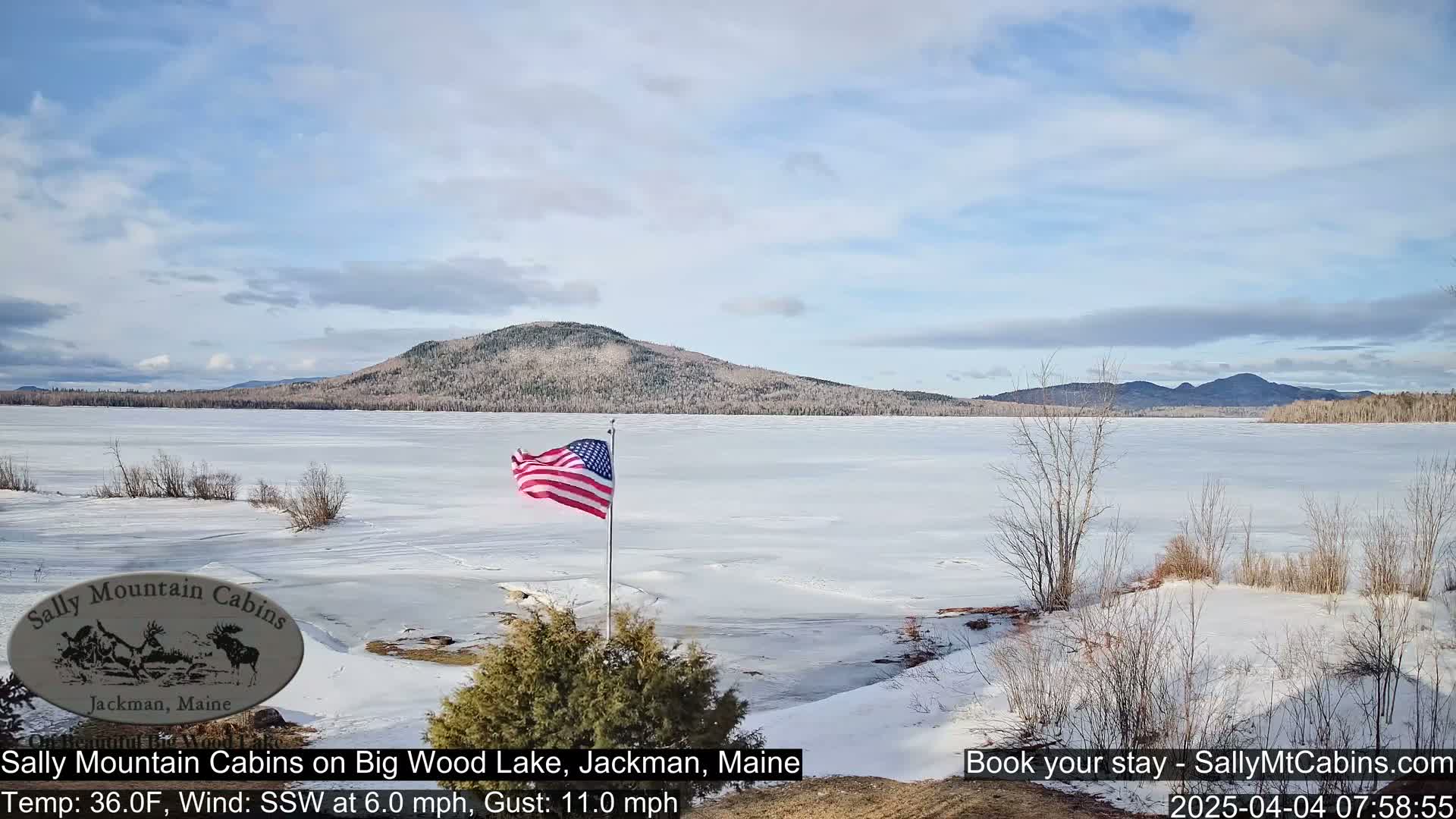 A partially frozen lake stretches out before a snow-covered shoreline, with a distant mountain range under a mostly clear, bright sky, and an American flag waving in the light breeze.
