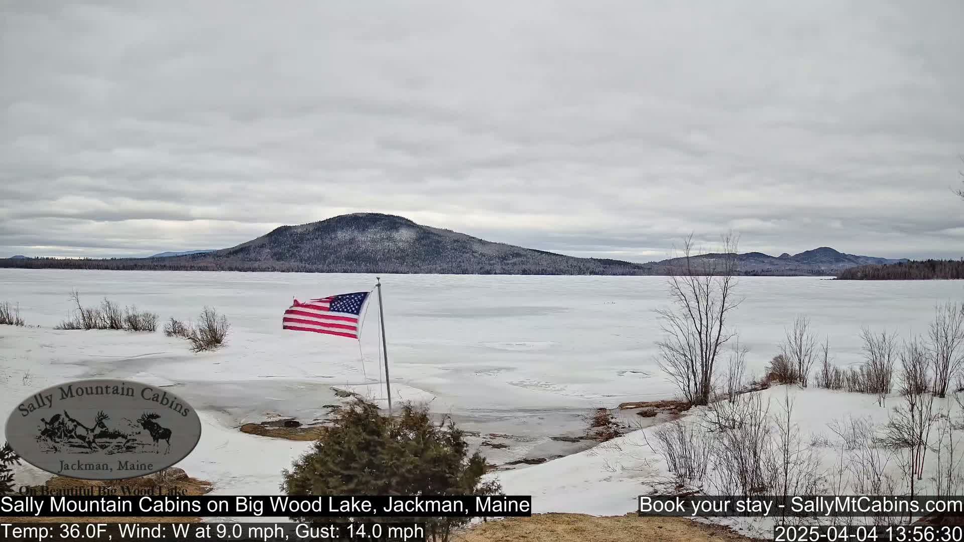 A partially frozen lake stretches out beneath an overcast sky, framed by snow-covered land and distant mountains, with an American flag waving in the wind.