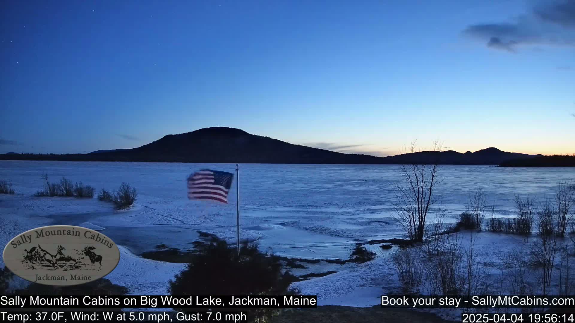 A partially frozen lake is seen at dusk under a clear sky, with a US flag waving in the wind from a lakeside pole, snow on the ground, and a dark mountain in the distance.