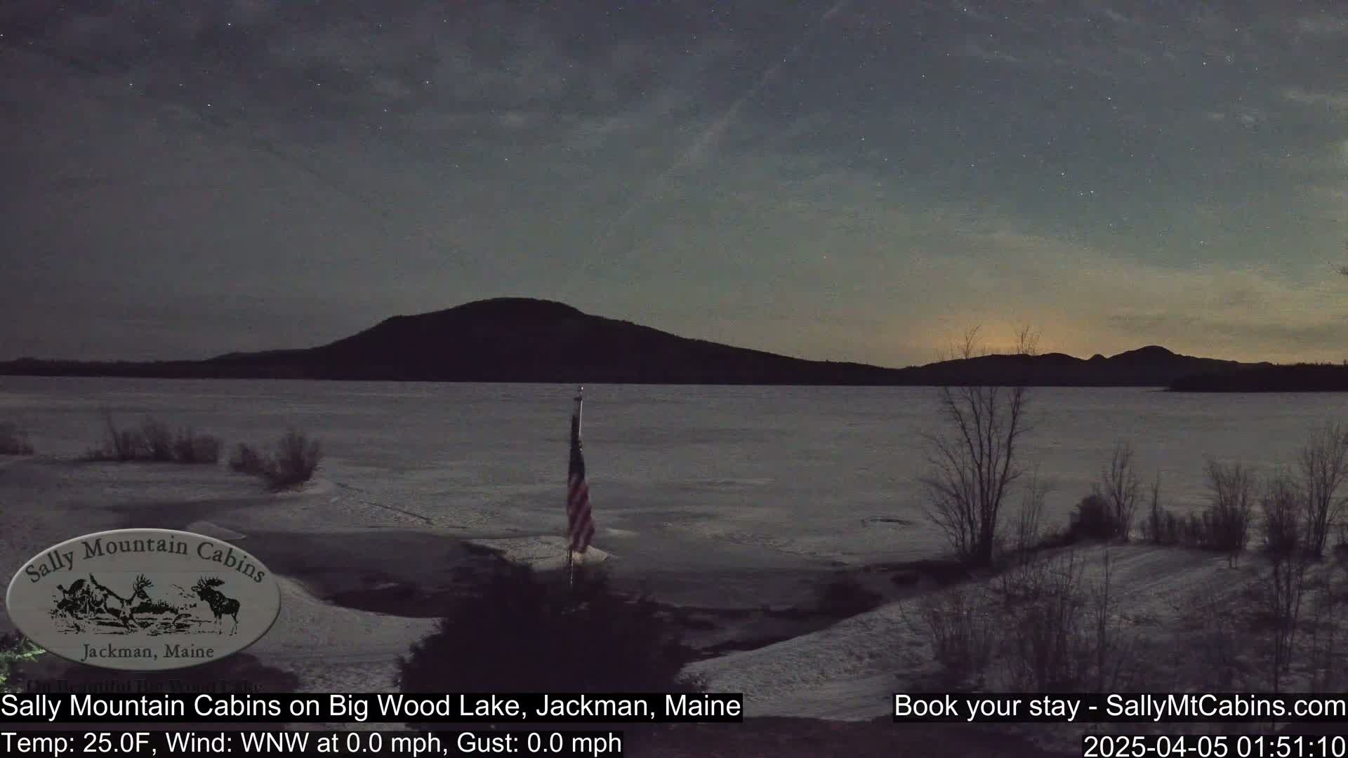 A partially frozen lake under a starry night sky, with a distant mountain range and a small American flag planted near the shore.