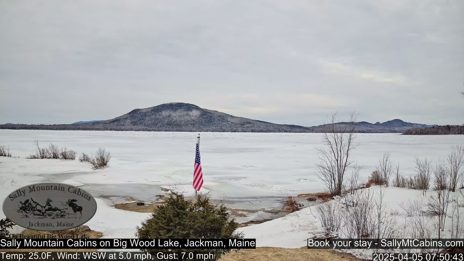 A snow-covered lake with partially melted ice, a distant mountain range, and an American flag on a flagpole are visible under an overcast sky.