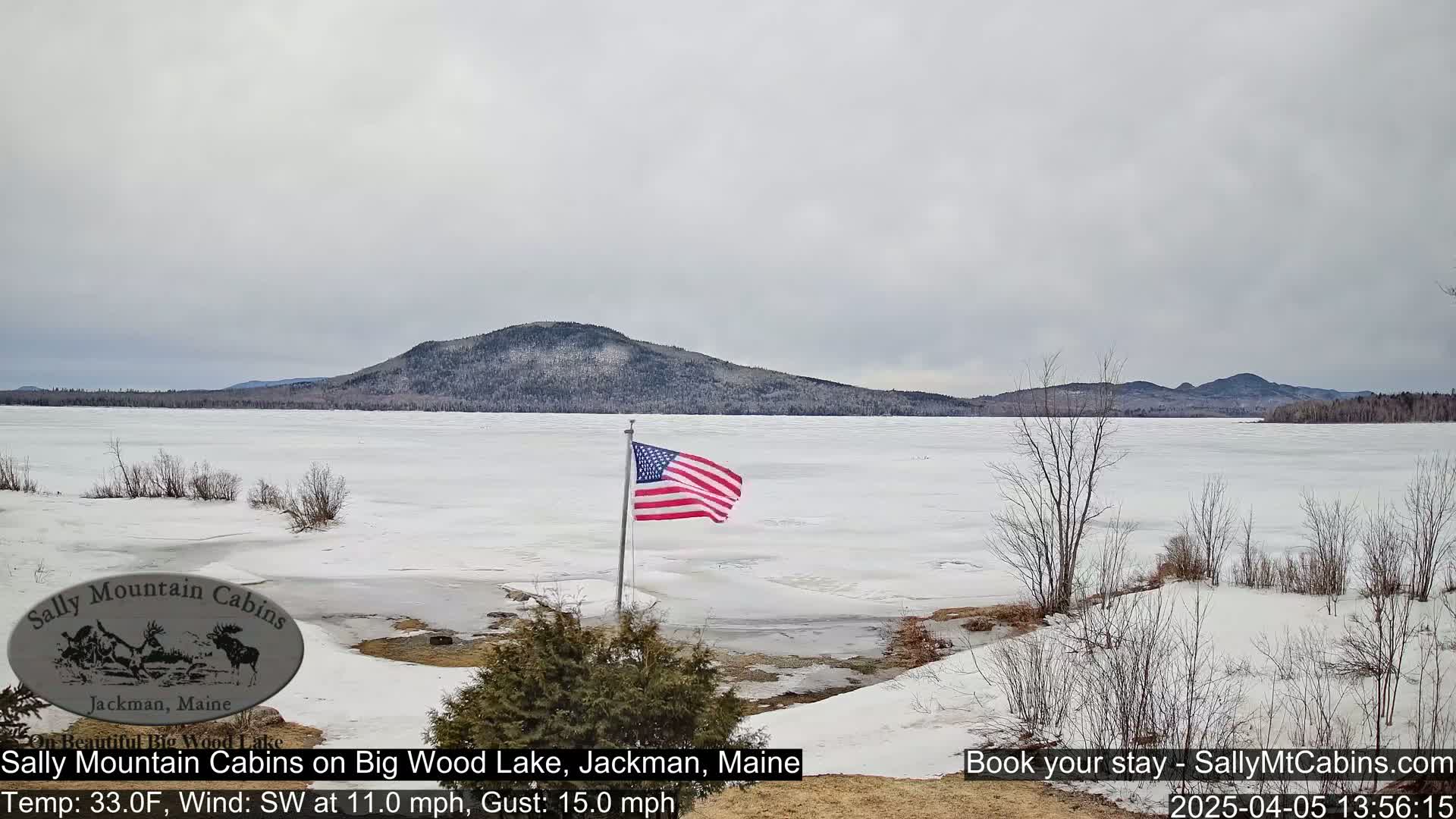 A partially frozen lake stretches out before snow-covered land, a partly cloudy sky overhead, and an American flag waving in the wind.