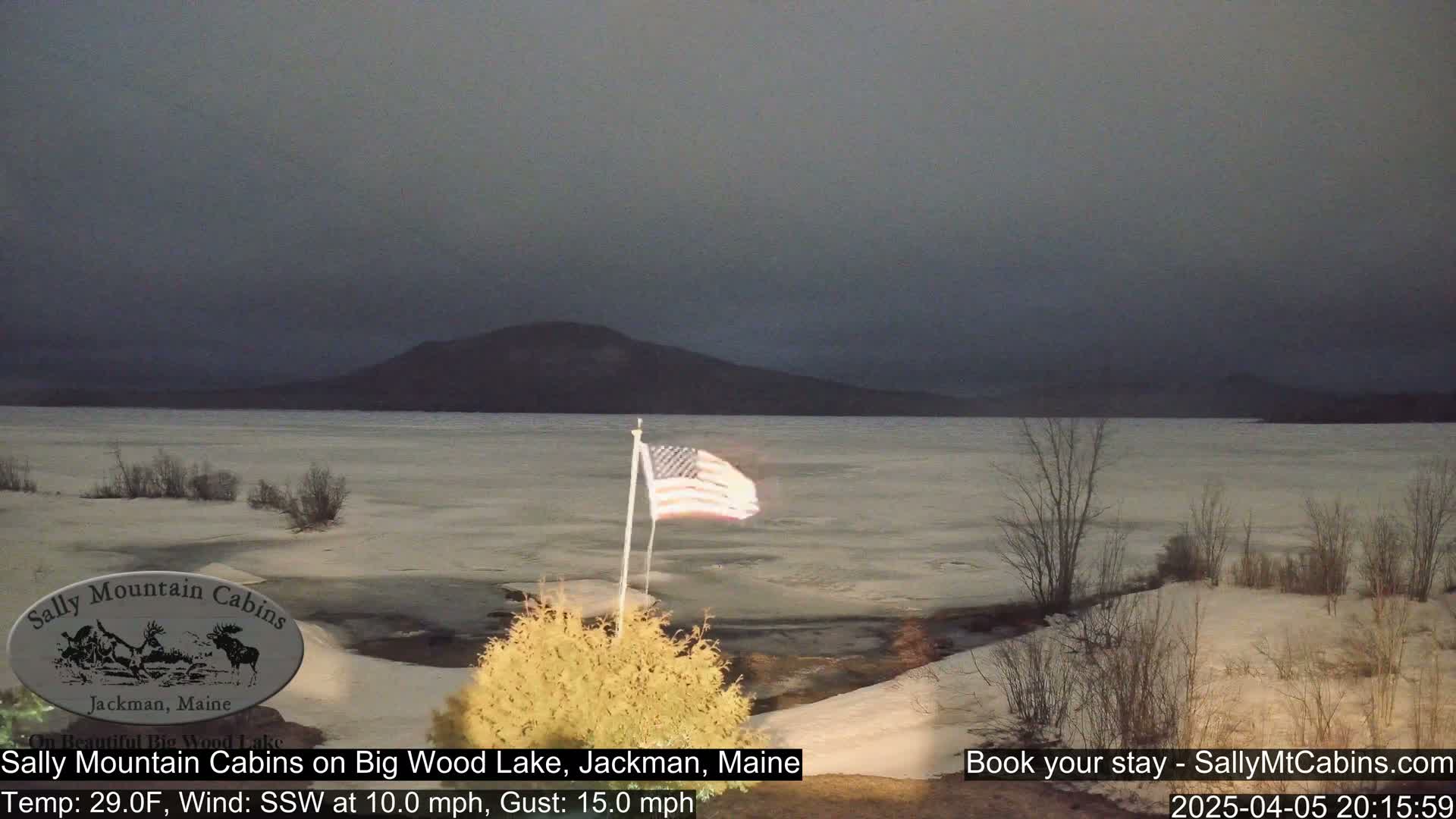 A partially frozen lake is visible under a dark, overcast sky, with a small American flag waving in the breeze near a snow-covered shoreline and distant mountains.