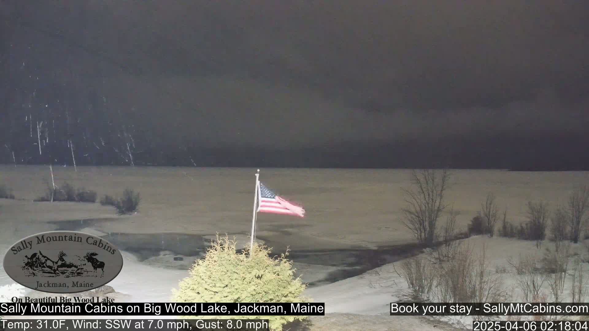 Snow falls over a partially frozen lake under a dark sky, with an American flag waving in the wind.
