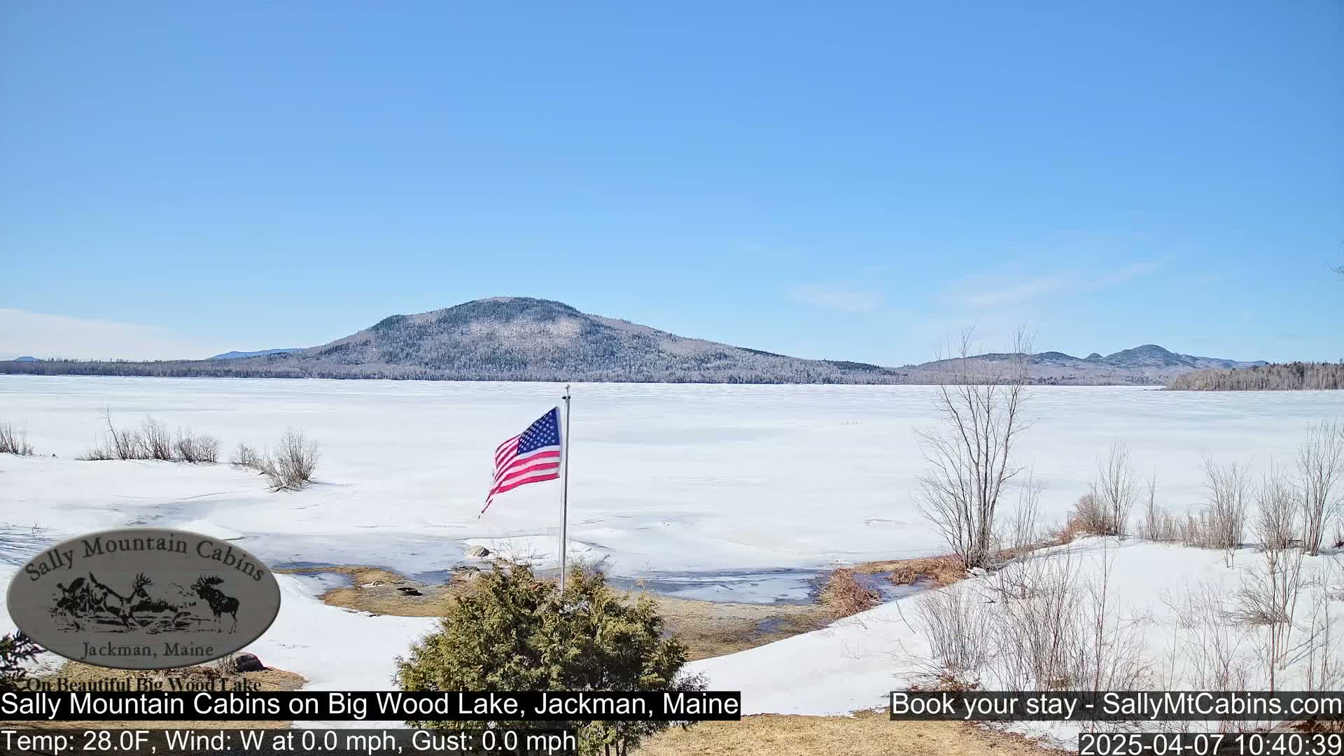 A snow-covered frozen lake stretches out before a partly wooded, snow-dusted mountain under a clear blue sky; an American flag stands in the foreground.