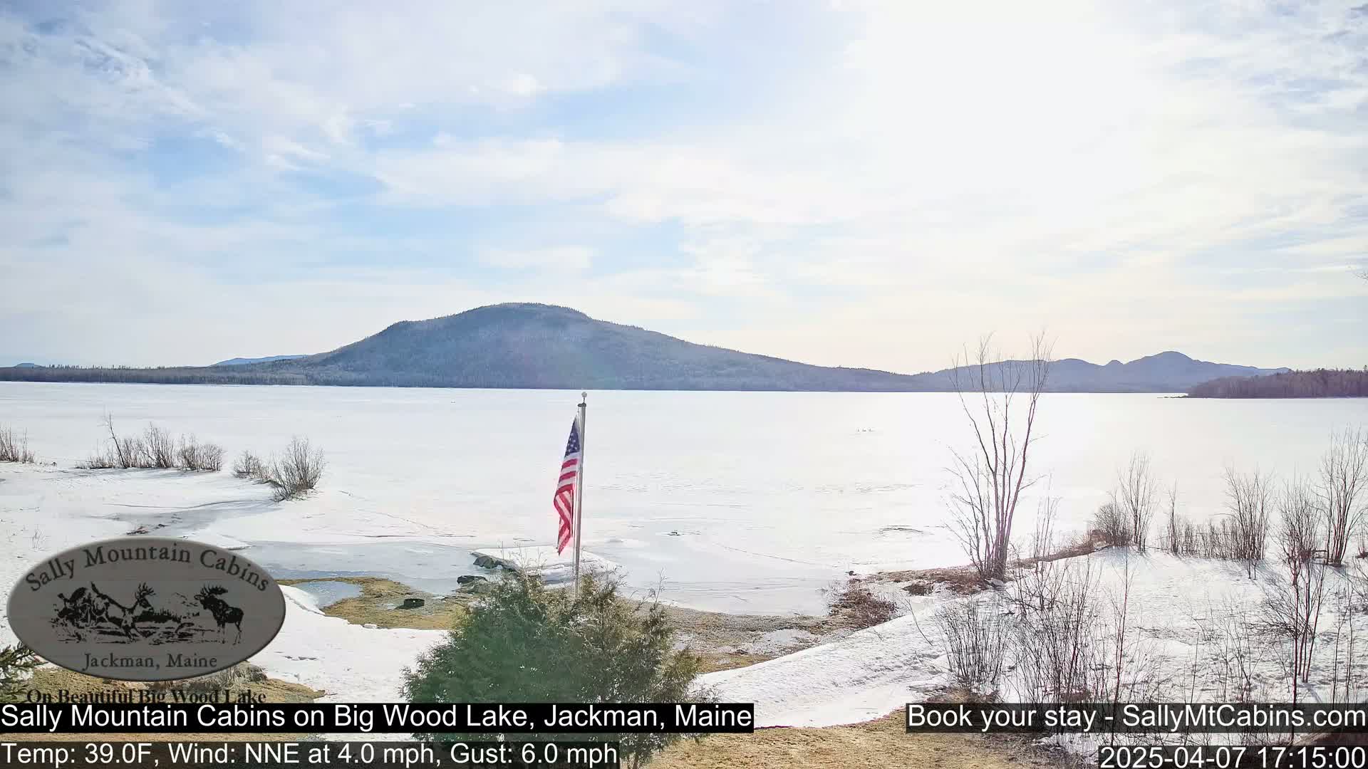 A partly frozen lake stretches toward snow-covered mountains under a mostly clear sky.