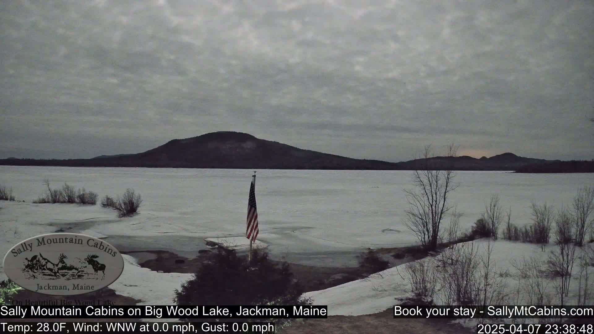 A partly frozen lake stretches out under a gray sky towards a distant, dark mountain range, with an American flag planted in the snow near the shoreline.
