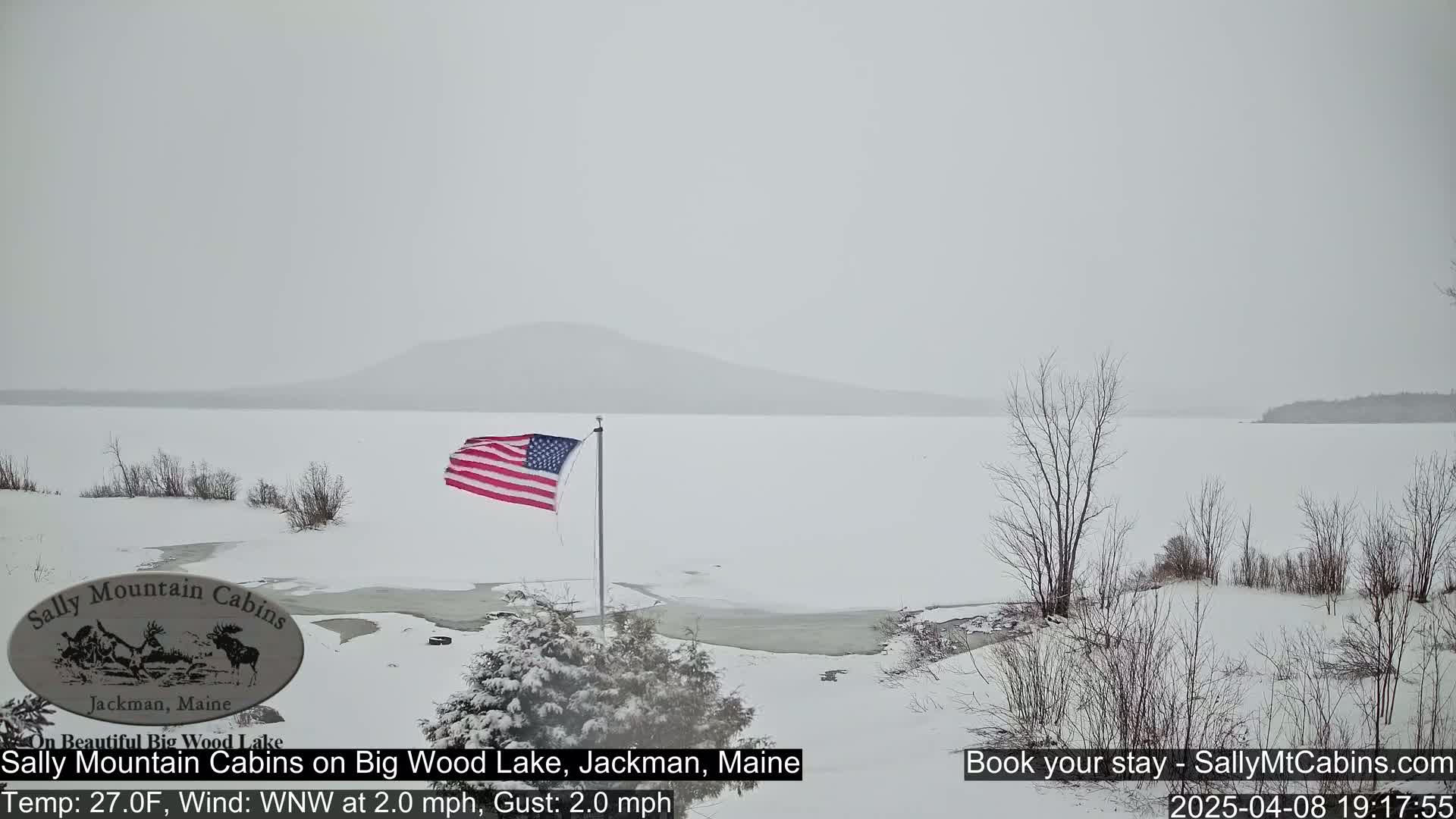 A snow-covered, partially frozen lake is seen from a snowy shoreline, with a US flag waving in the wind against a hazy, snowy background.