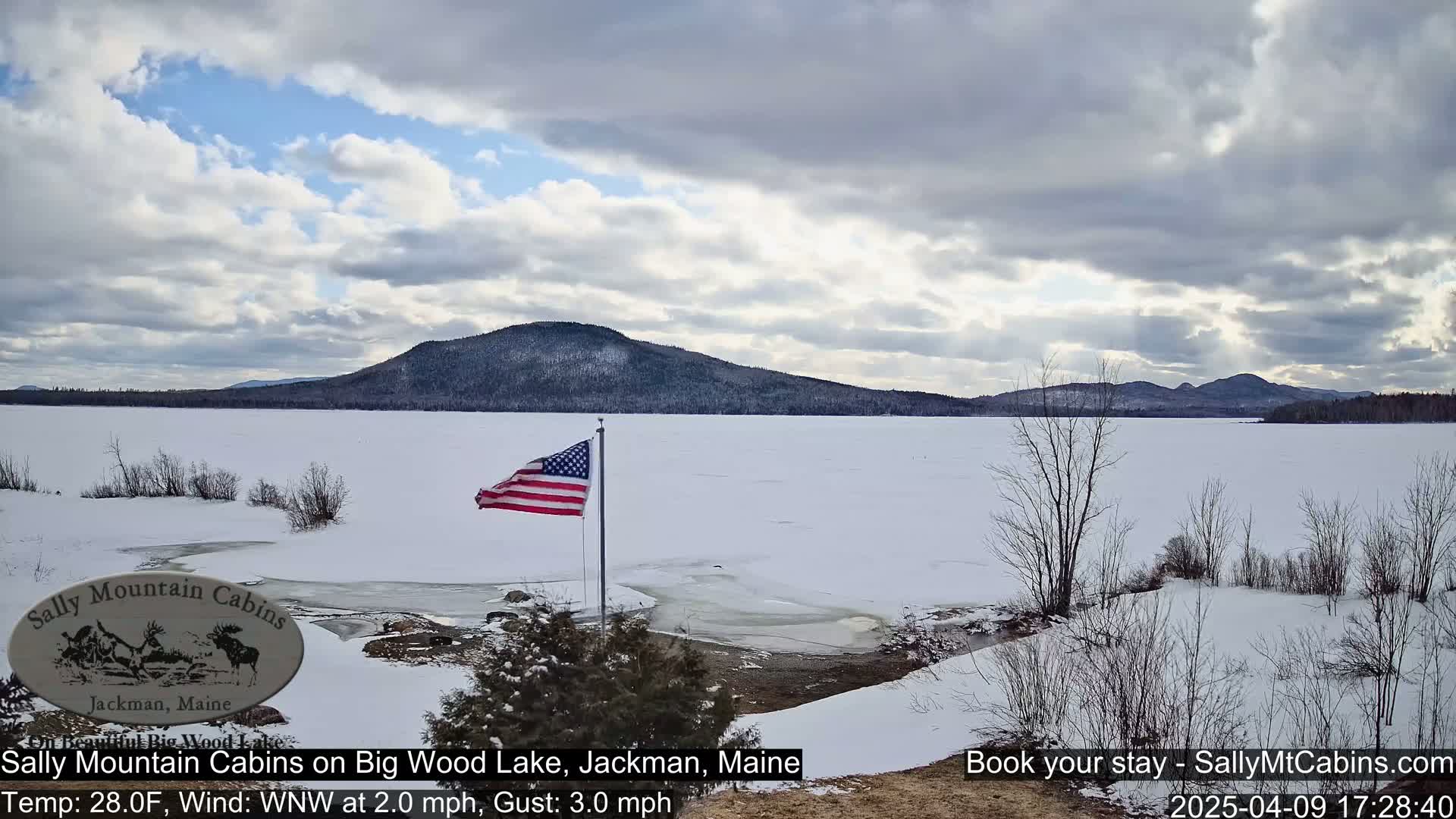 A snow-covered frozen lake stretches to a distant, wooded mountain range under a partly cloudy sky.  An American flag stands near the shore.