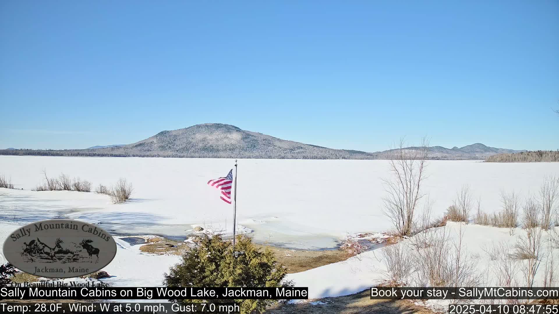 A snow-covered lake stretches to a snow-dusted mountain range under a clear blue sky, with an American flag waving in the foreground.