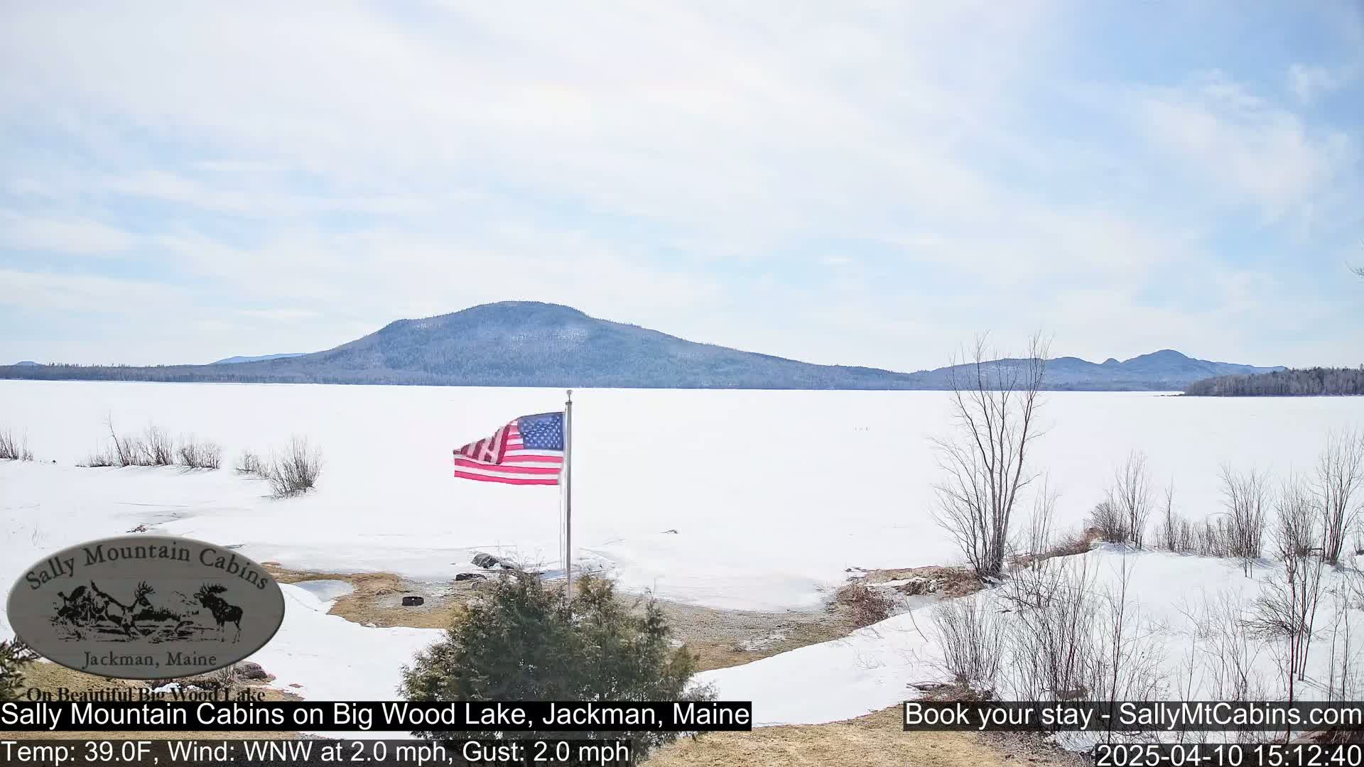 A frozen lake stretches to a distant mountain range under a mostly clear sky, with an American flag waving in the foreground.