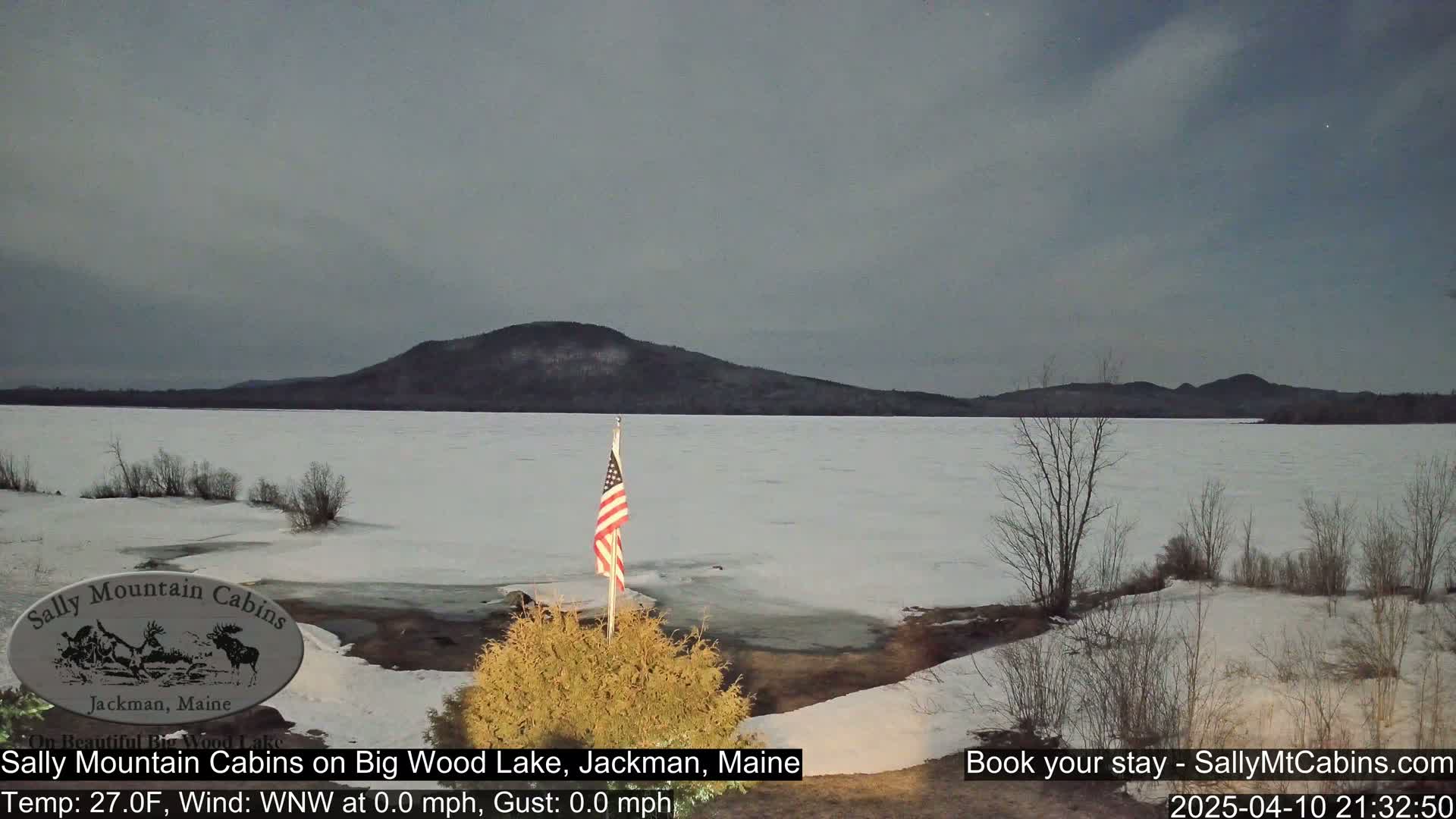 A partially frozen lake stretches to a snow-dusted mountain under a gray, twilight sky; an American flag stands in the foreground.