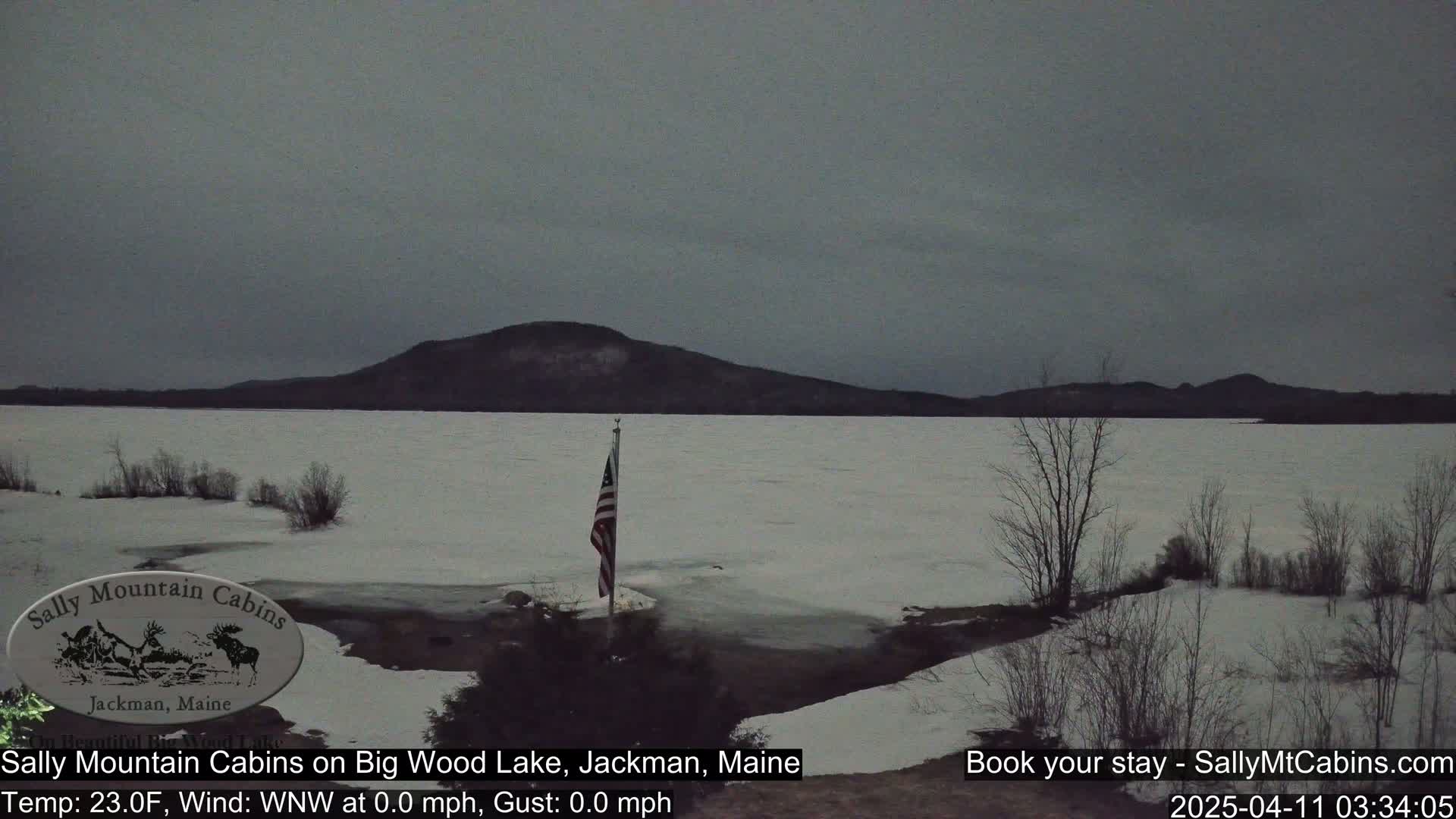 A partly frozen lake is visible under a gray sky, with a US flag standing on the shoreline and snow-covered land in the foreground and distant hills beyond the lake.