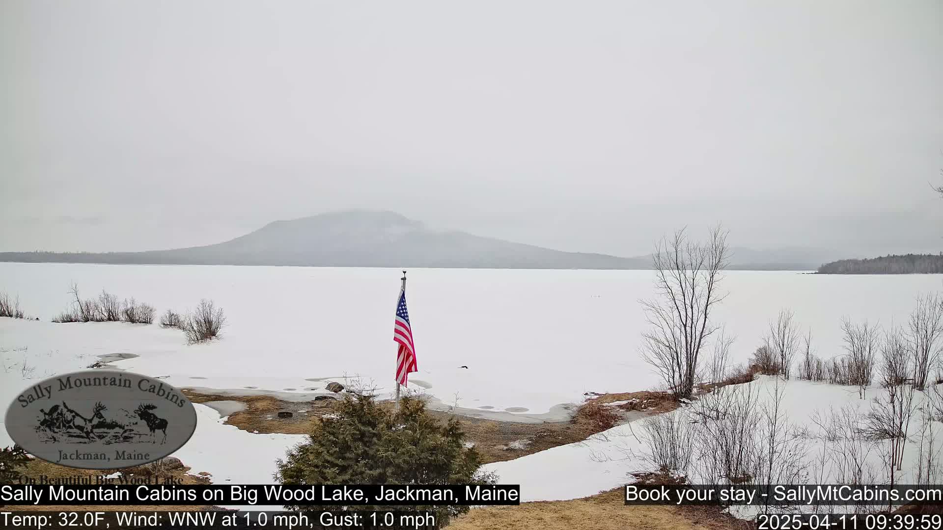 A snow-covered lake with a distant mountain range under an overcast sky, featuring a US flag in the foreground.