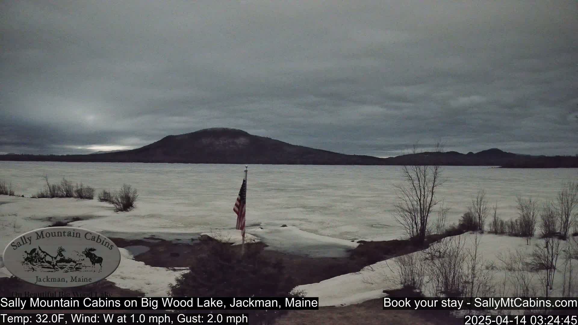 A partially frozen lake under a gray sky, with a distant mountain range and an American flag on a flagpole in the foreground.
