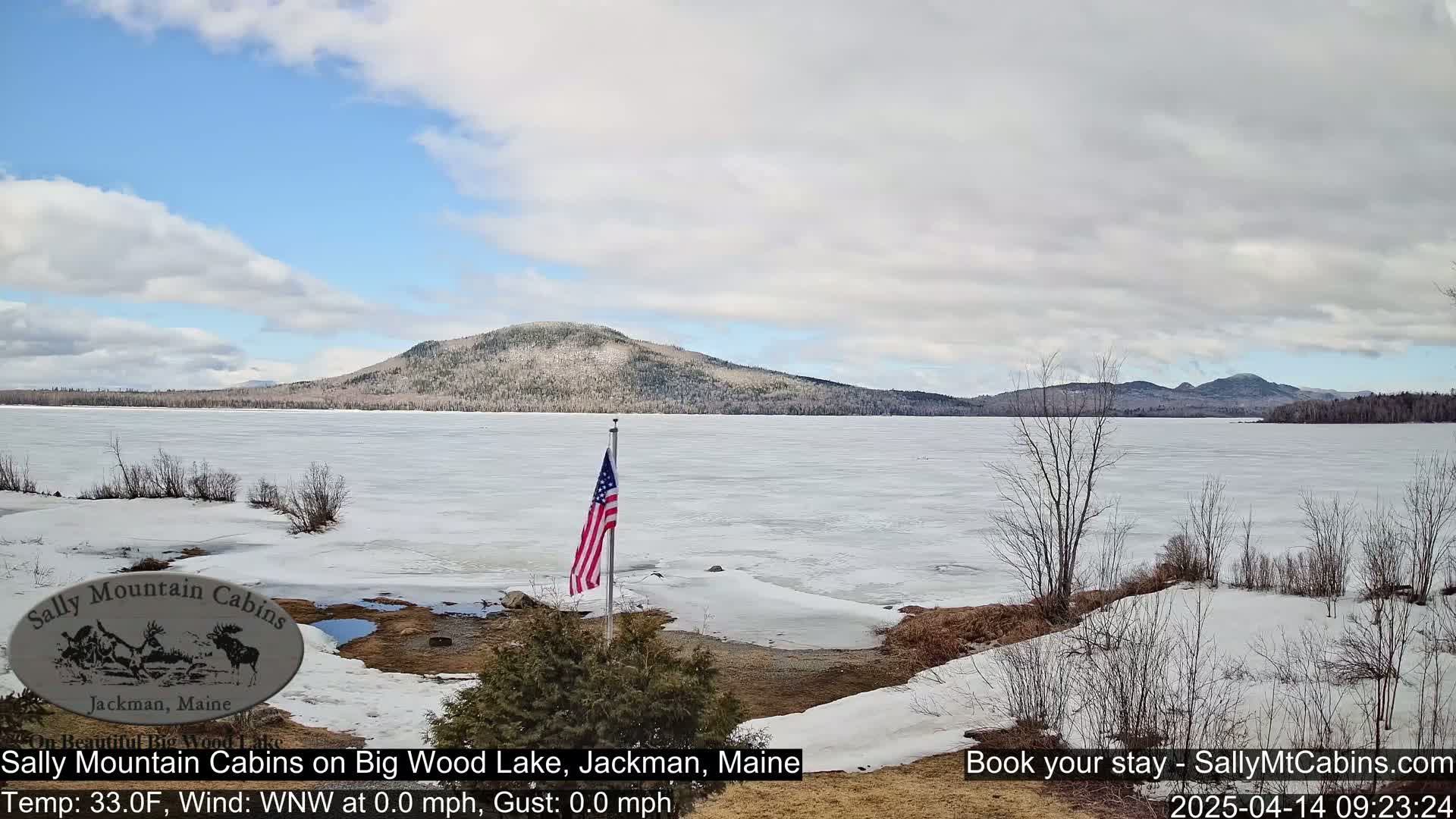 A partly cloudy day shows a mostly frozen lake with a snow-covered shoreline, a forested hill in the background, and an American flag on a pole in the foreground.