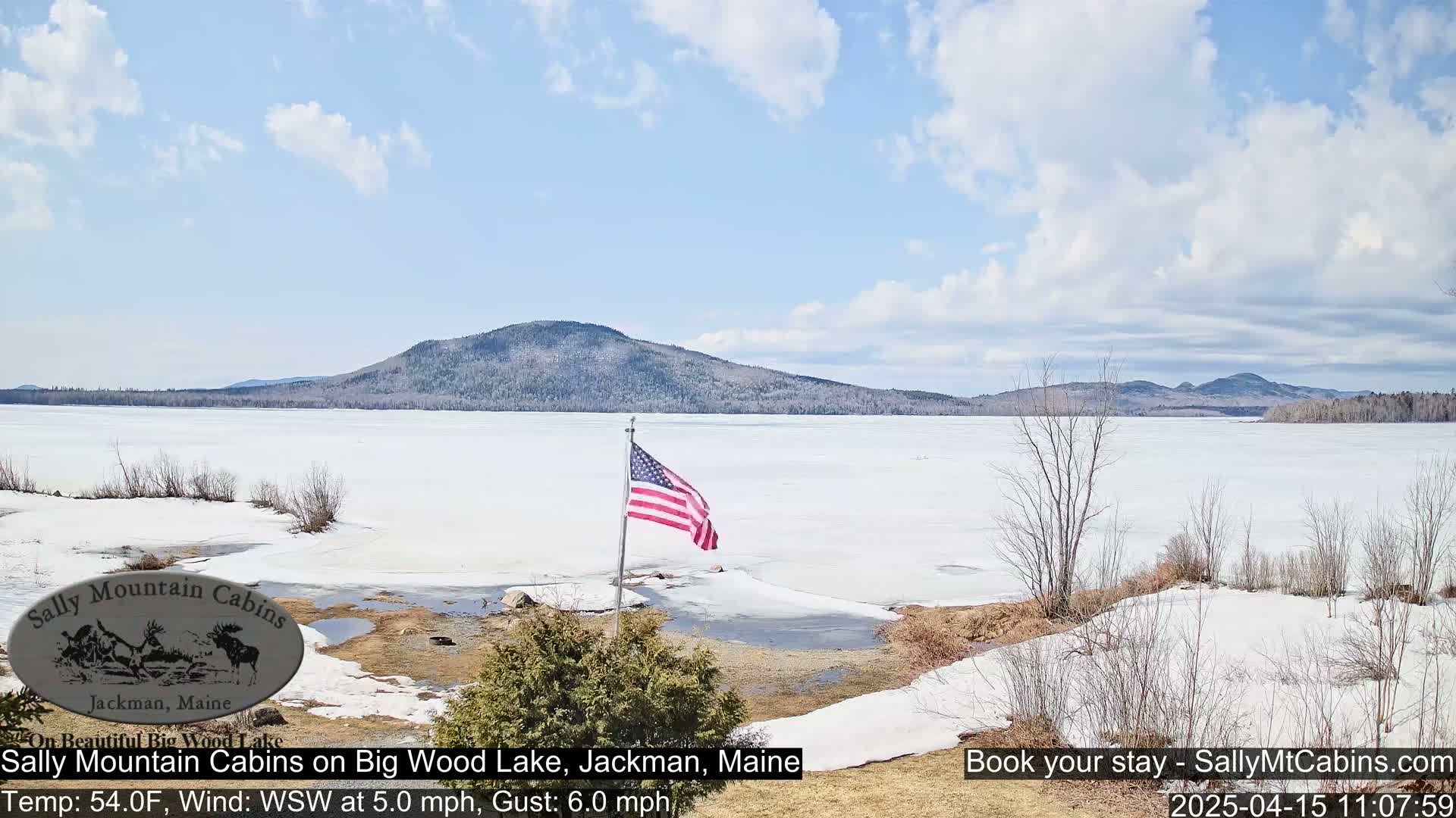 A partially frozen lake with a distant mountain range under a partly cloudy sky, and an American flag is displayed near the lake's edge.
