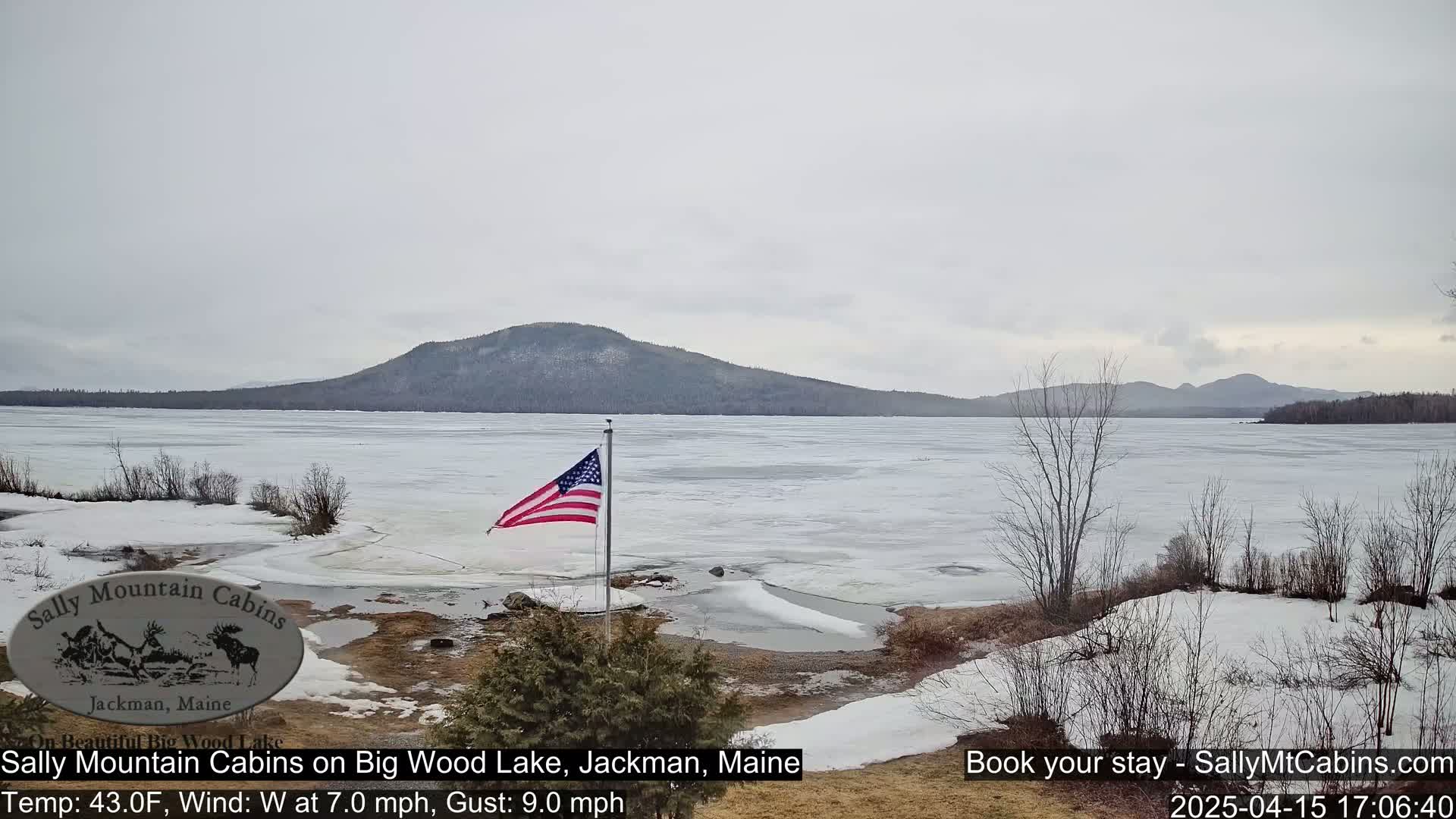 A partially frozen lake stretches towards a mountain range under an overcast sky, with an American flag waving in the foreground.