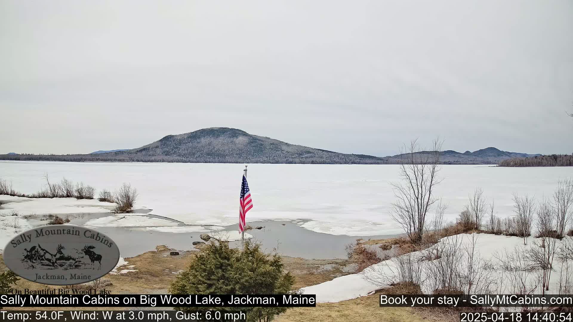A partially frozen lake stretches towards snow-covered hills under a cloudy sky, with an American flag planted on the lakeshore.