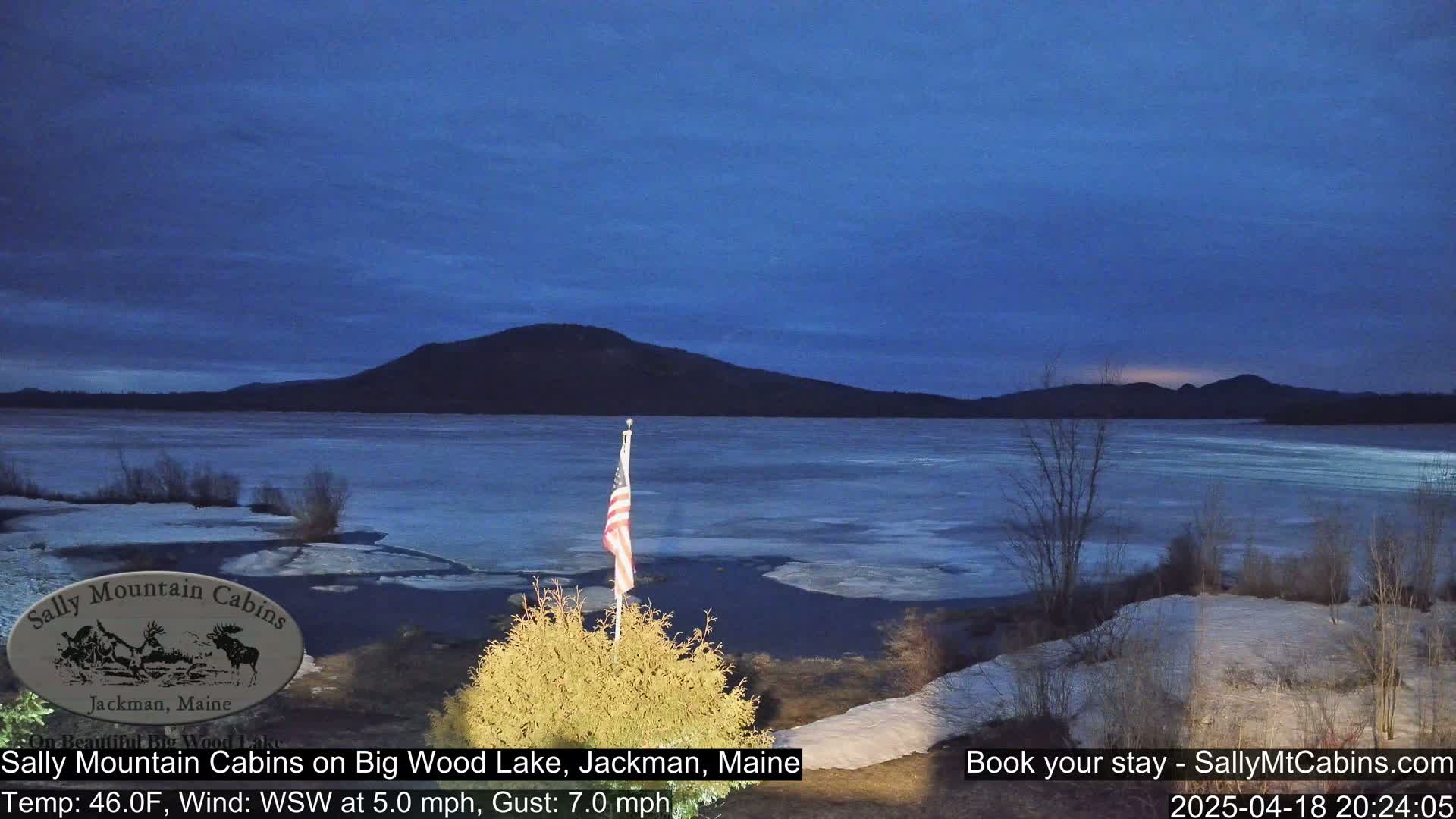 A partially frozen lake is seen at dusk under a twilight blue sky, with a small American flag in the foreground.