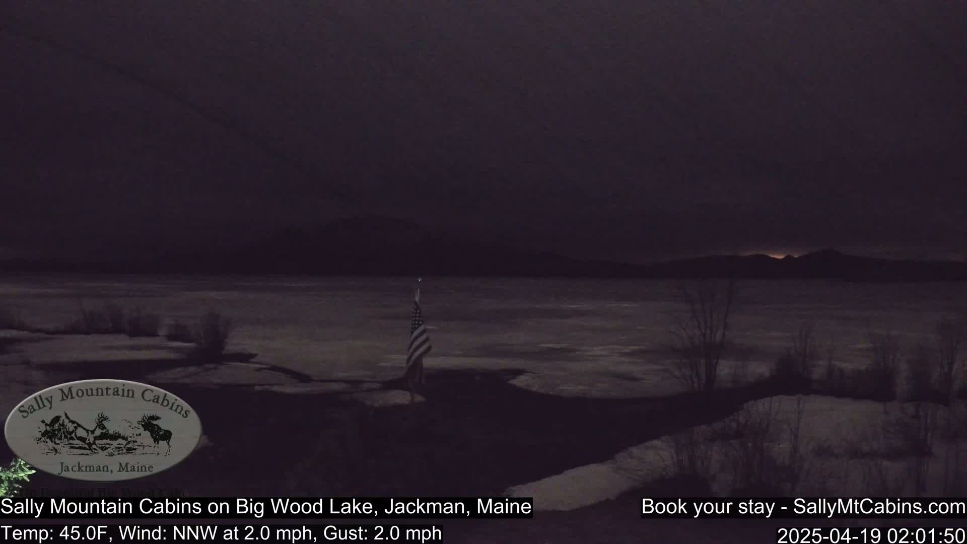 A partially frozen lake is visible under a dark, nighttime sky, with a flagpole and an American flag standing near the shoreline.