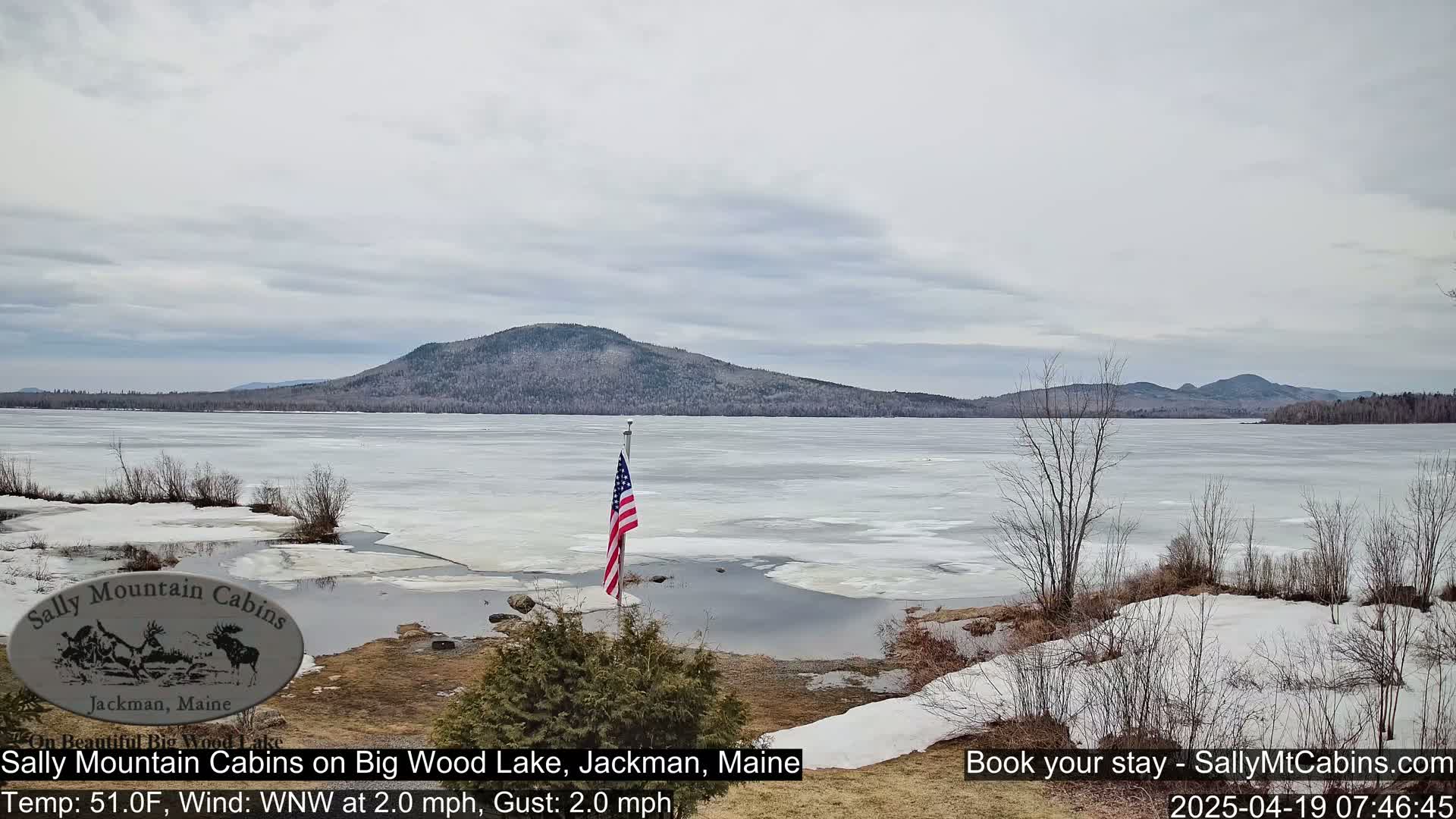 A partly frozen lake stretches to a distant mountain range under a cloudy sky.