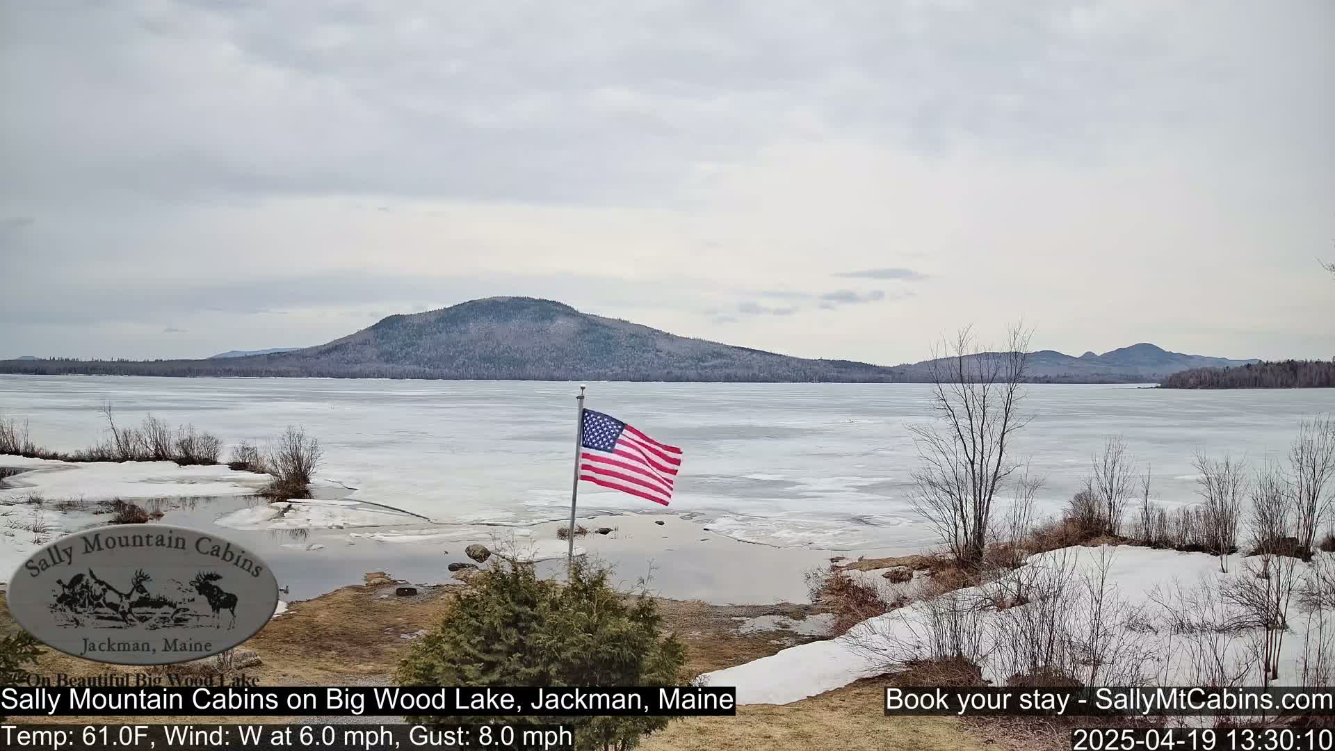 A partially frozen lake is seen under a cloudy sky, with a gently waving American flag in the foreground and mountains in the background.