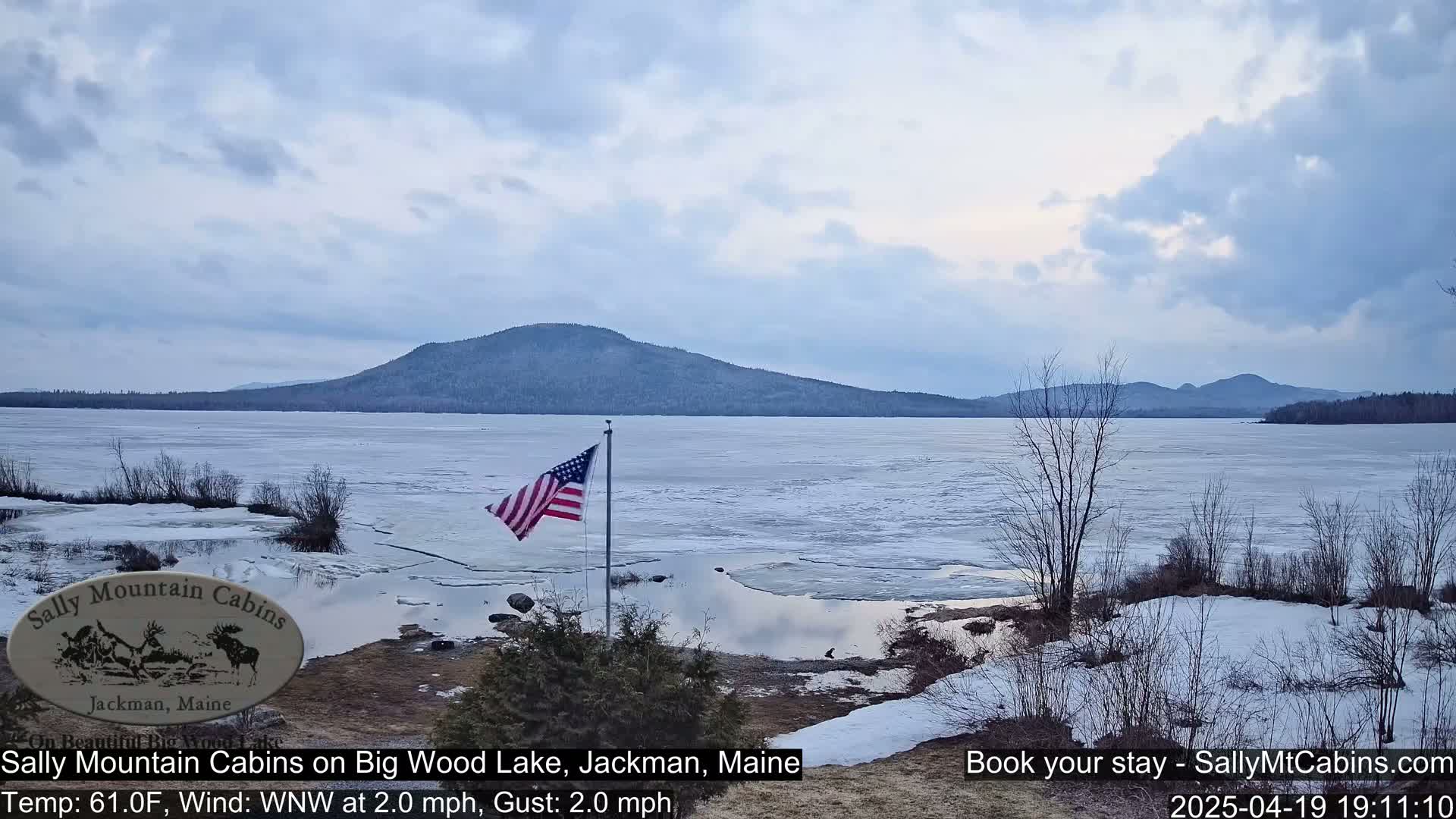 A partly cloudy sky overlooks a mostly frozen lake with melting patches of ice near the shore, mountains in the background, and an American flag waving in the breeze.