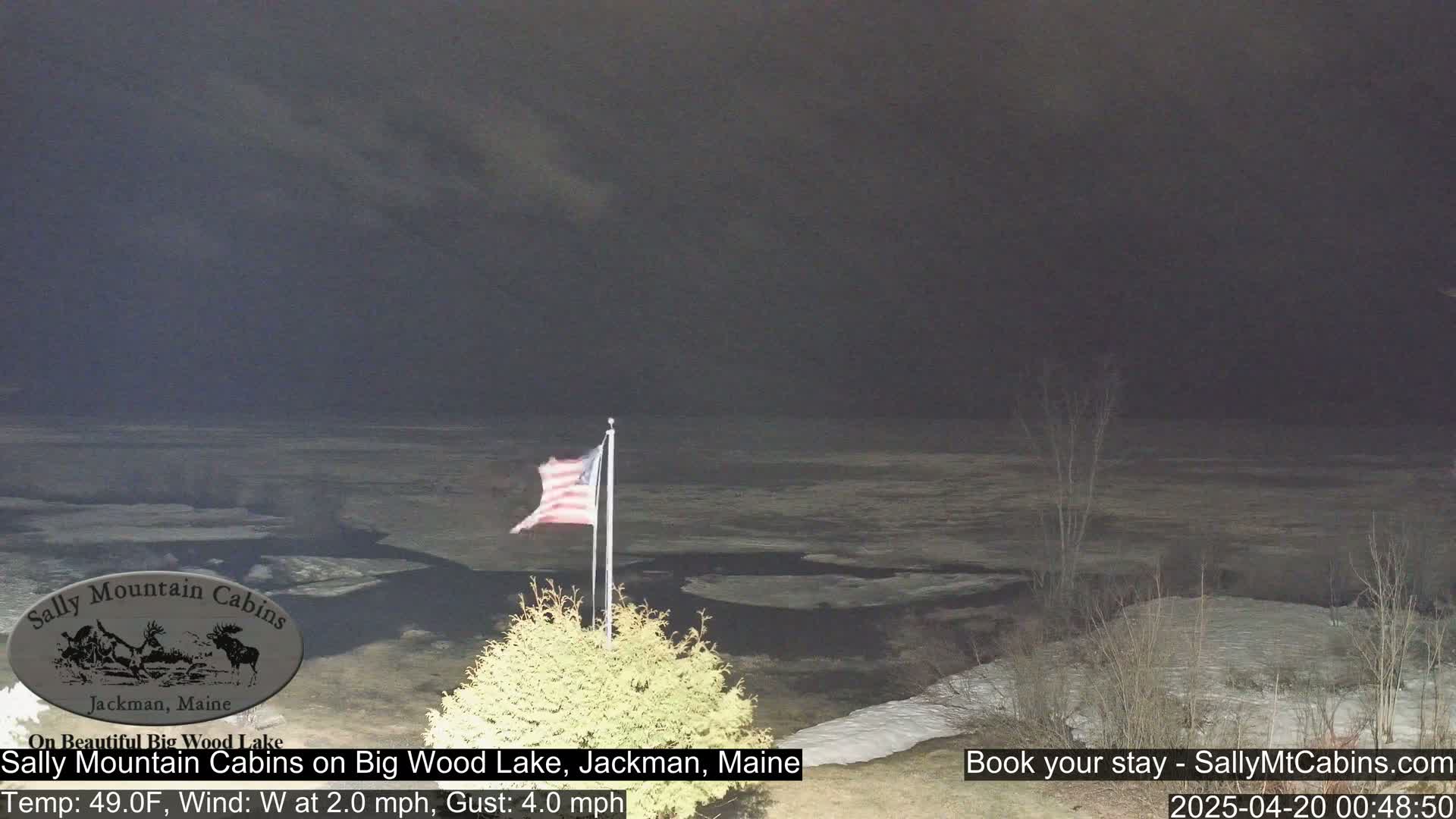 A partially frozen lake is visible at night under a dark, overcast sky, with an American flag waving gently in the foreground.