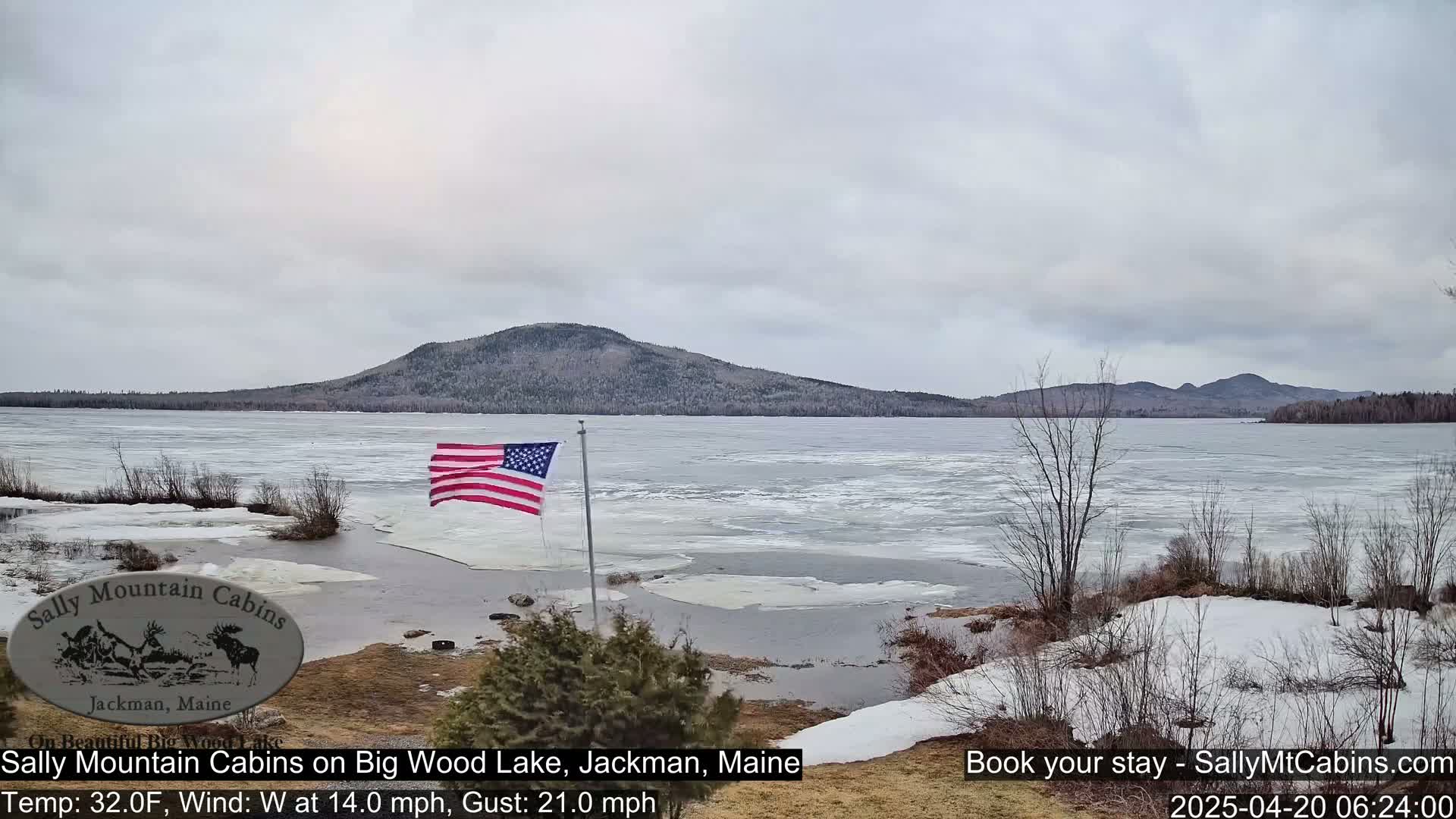 A partially frozen lake with a hilly background under an overcast sky, and an American flag waving near the shoreline.