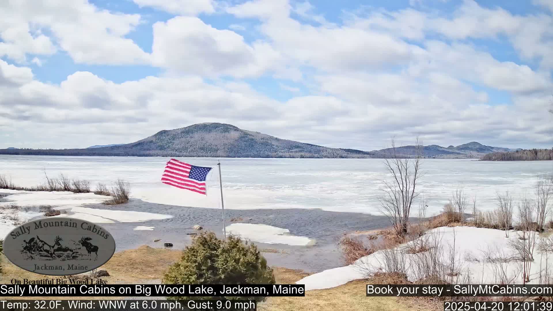 A partially frozen lake, with a partly cloudy sky, is seen from a lakeside vantage point, where an American flag flies near the shore.