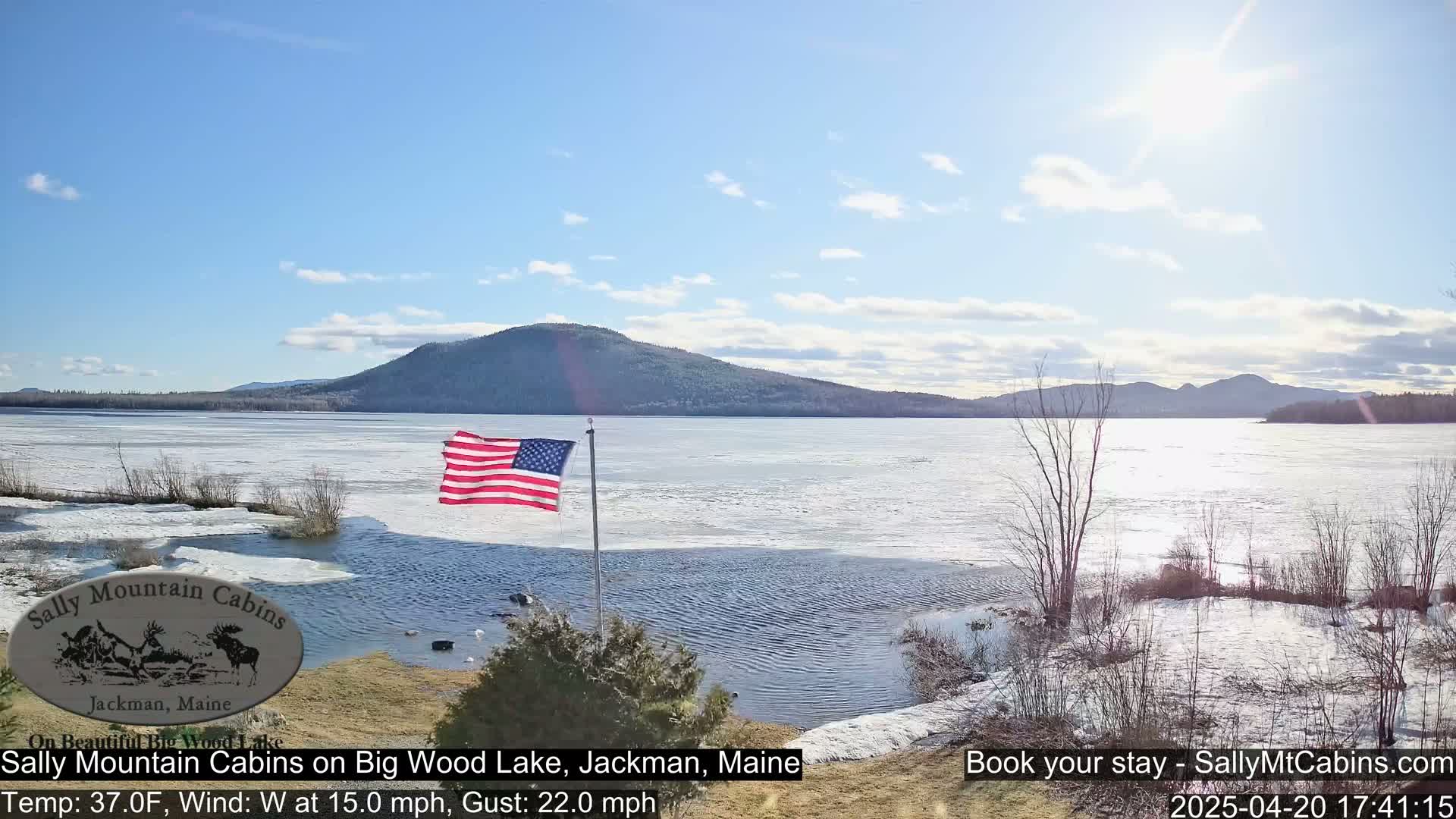 An American flag flies near a partially frozen lake under a mostly sunny sky, with snow and bare trees on the shoreline and mountains in the background.