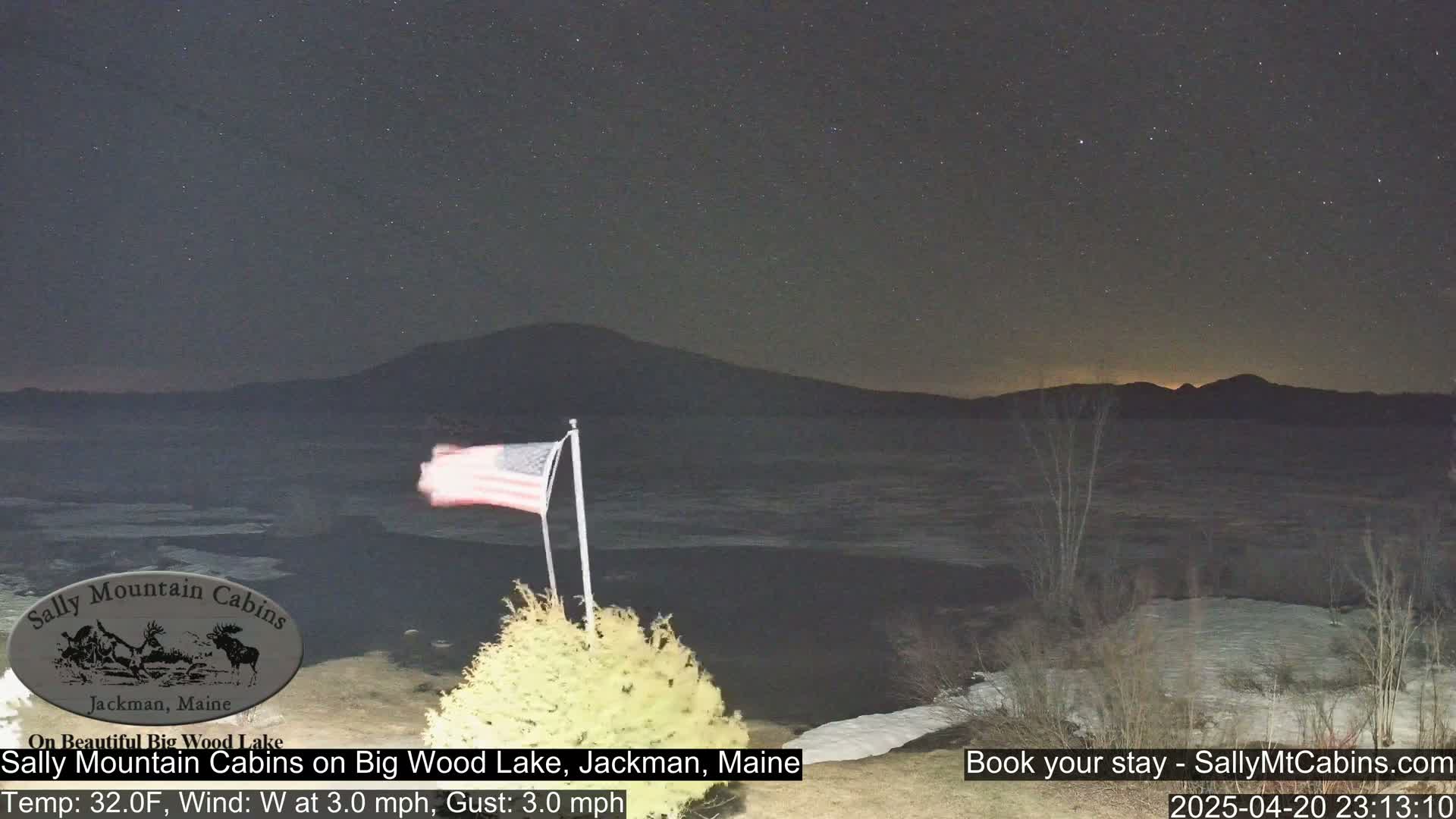 A partly frozen lake is seen under a starry night sky, with a distant mountain range and an American flag in the foreground.