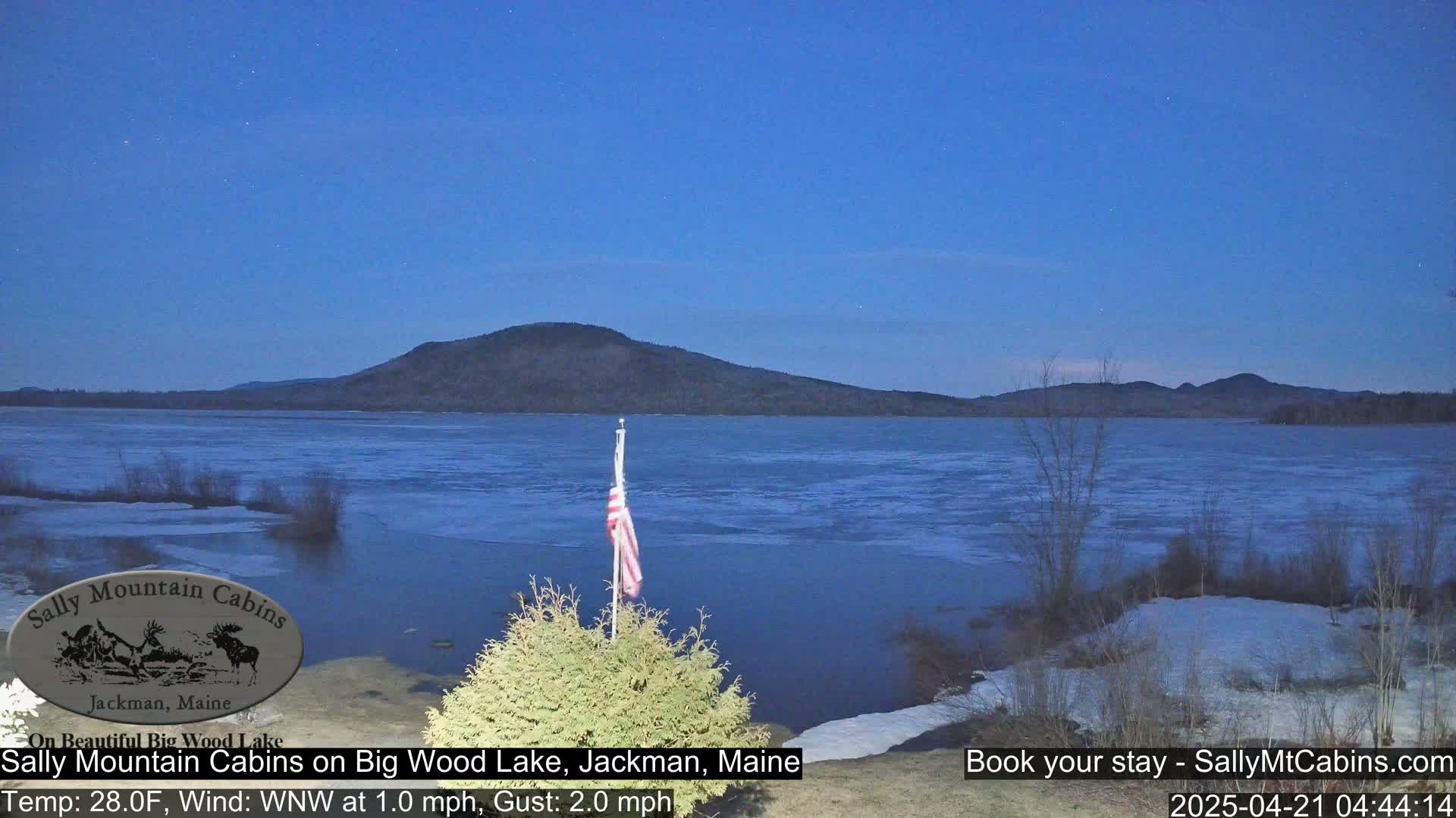 A partly frozen lake is seen at twilight under a clear, dark blue sky, with a flagpole and small evergreen bush in the foreground and a mountain in the background.