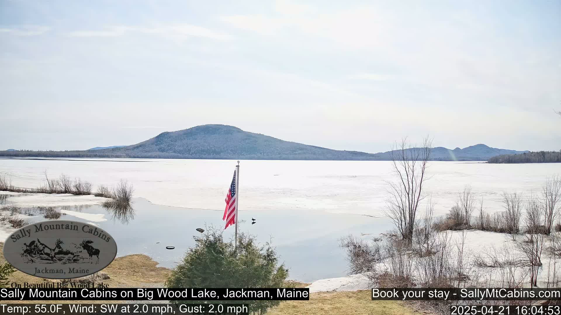 A partly frozen lake stretches to a distant mountain range under a mostly clear sky.