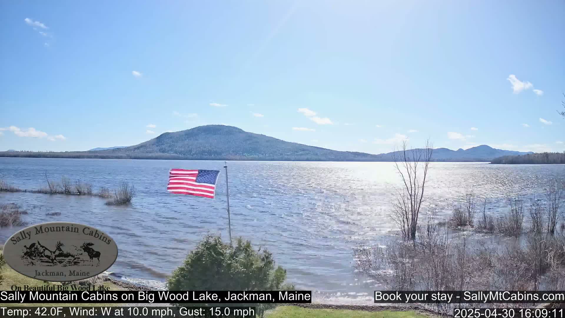 An American flag flies near a lake under a mostly clear sky, with a mountain in the distance and a slight chop on the water.