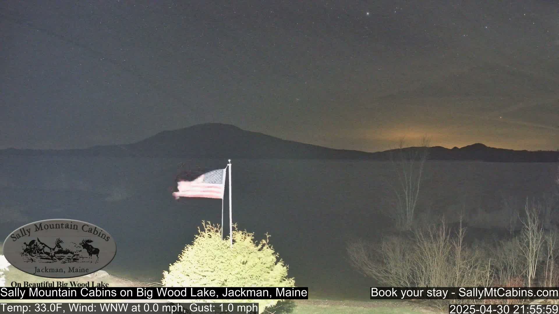 A nighttime view of a calm lake with a mountain range in the background under a starry sky, with an American flag waving gently in the foreground.