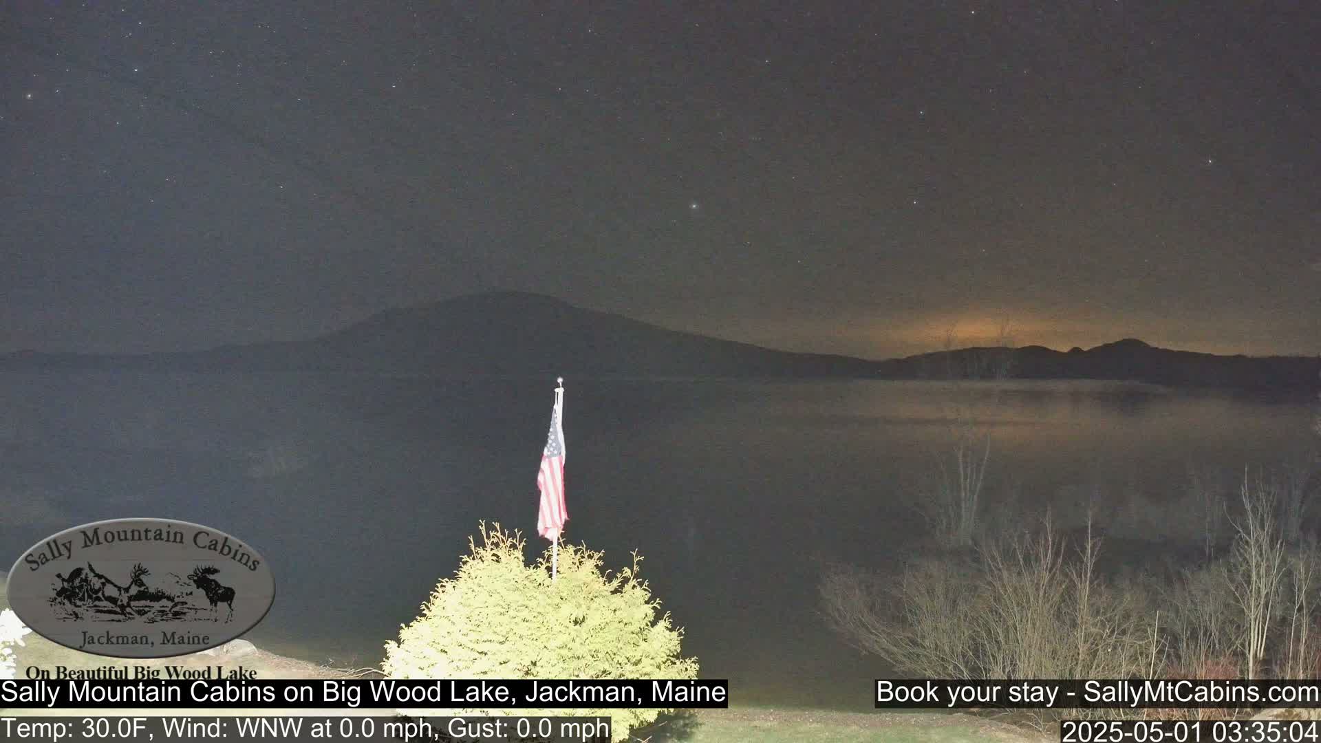 A calm, starry night scene shows a lake reflecting the sky, mountains in the distance, and an American flag on a pole in the foreground.